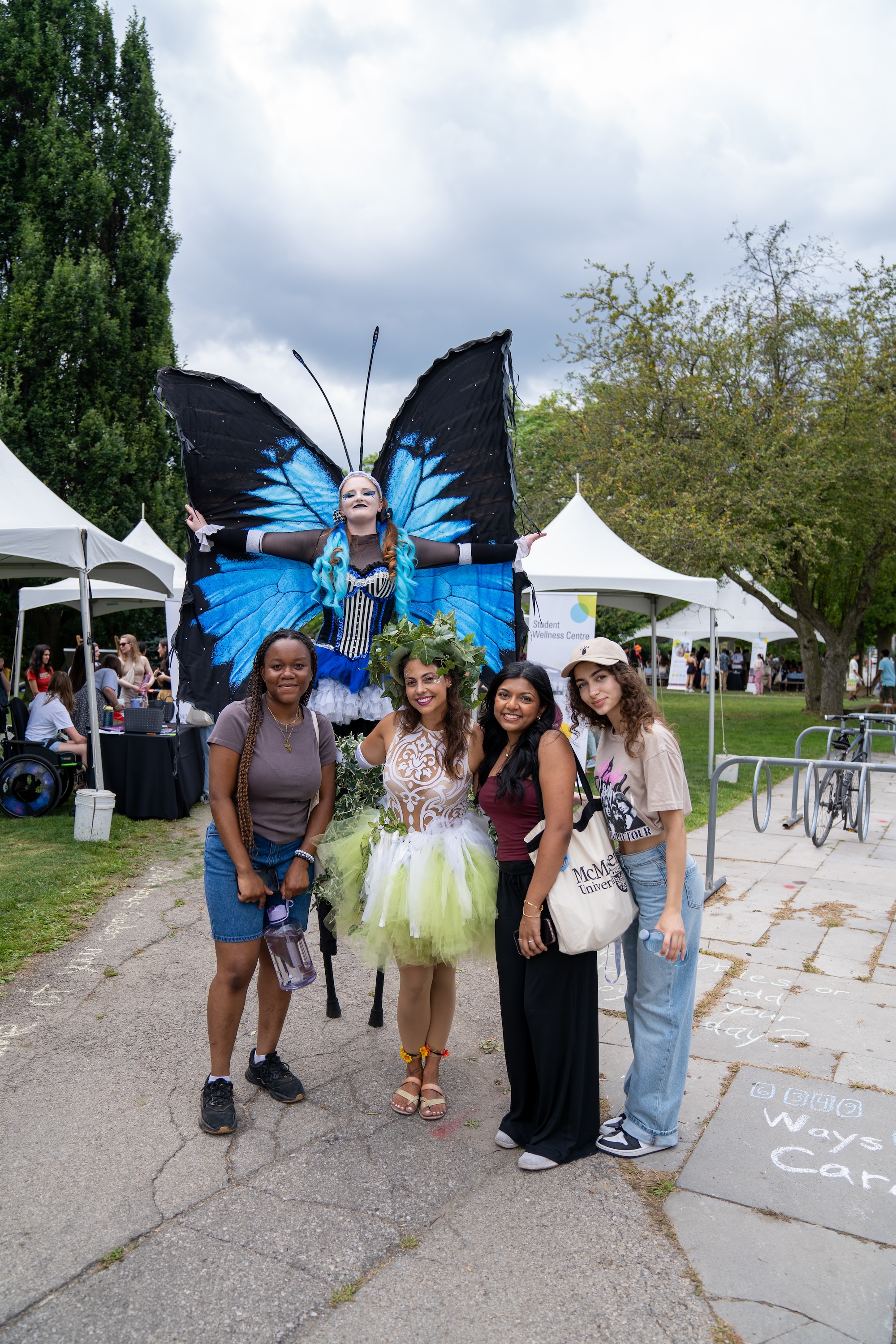 A stiltwalker with enormous butterfly wings stands with students and a person dressed like a fairy, outdoors on campus.