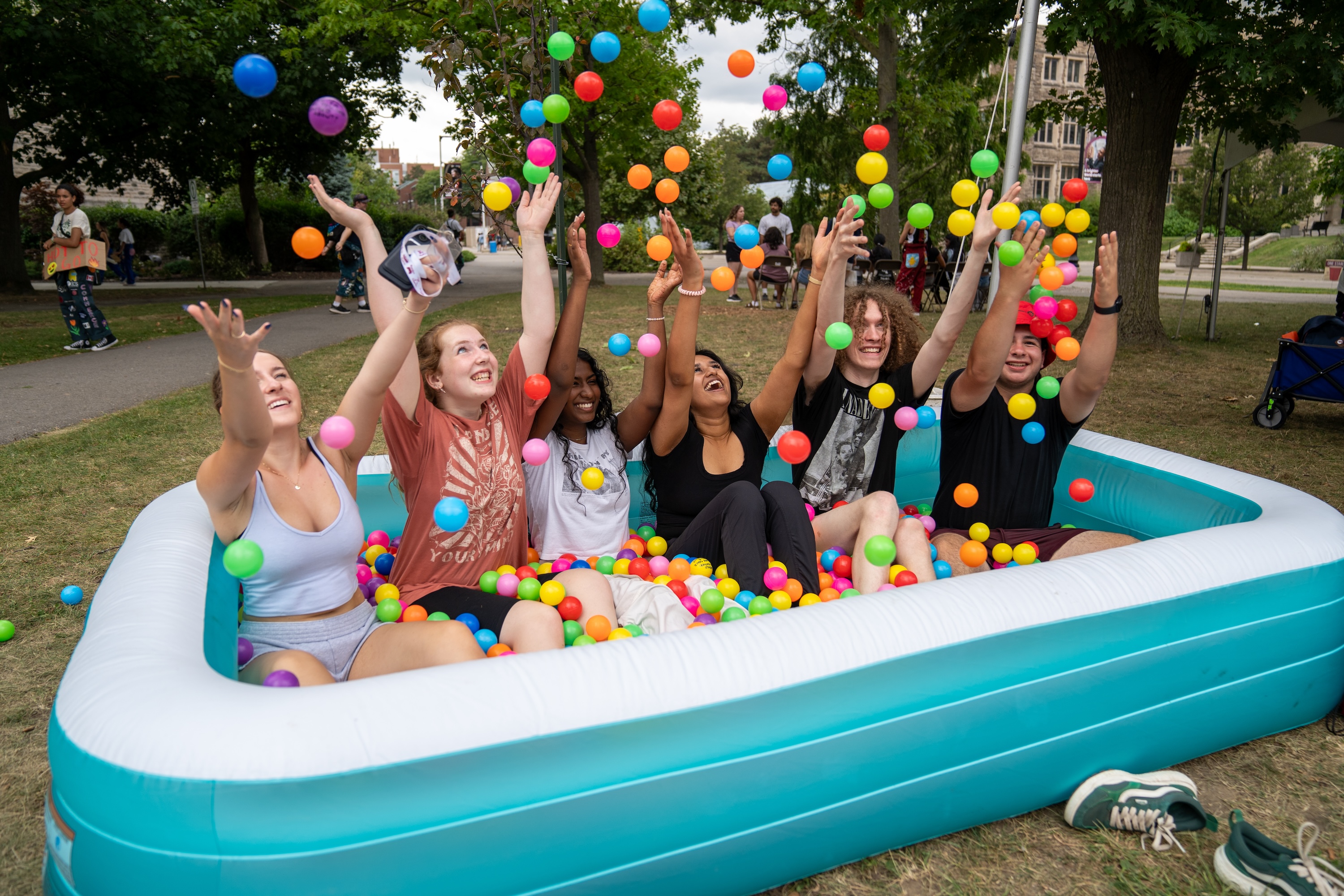 Five young people sit in an inflatable wading pool full of colourful plastic balls with their hands up in the air as more plastic rain down on them.