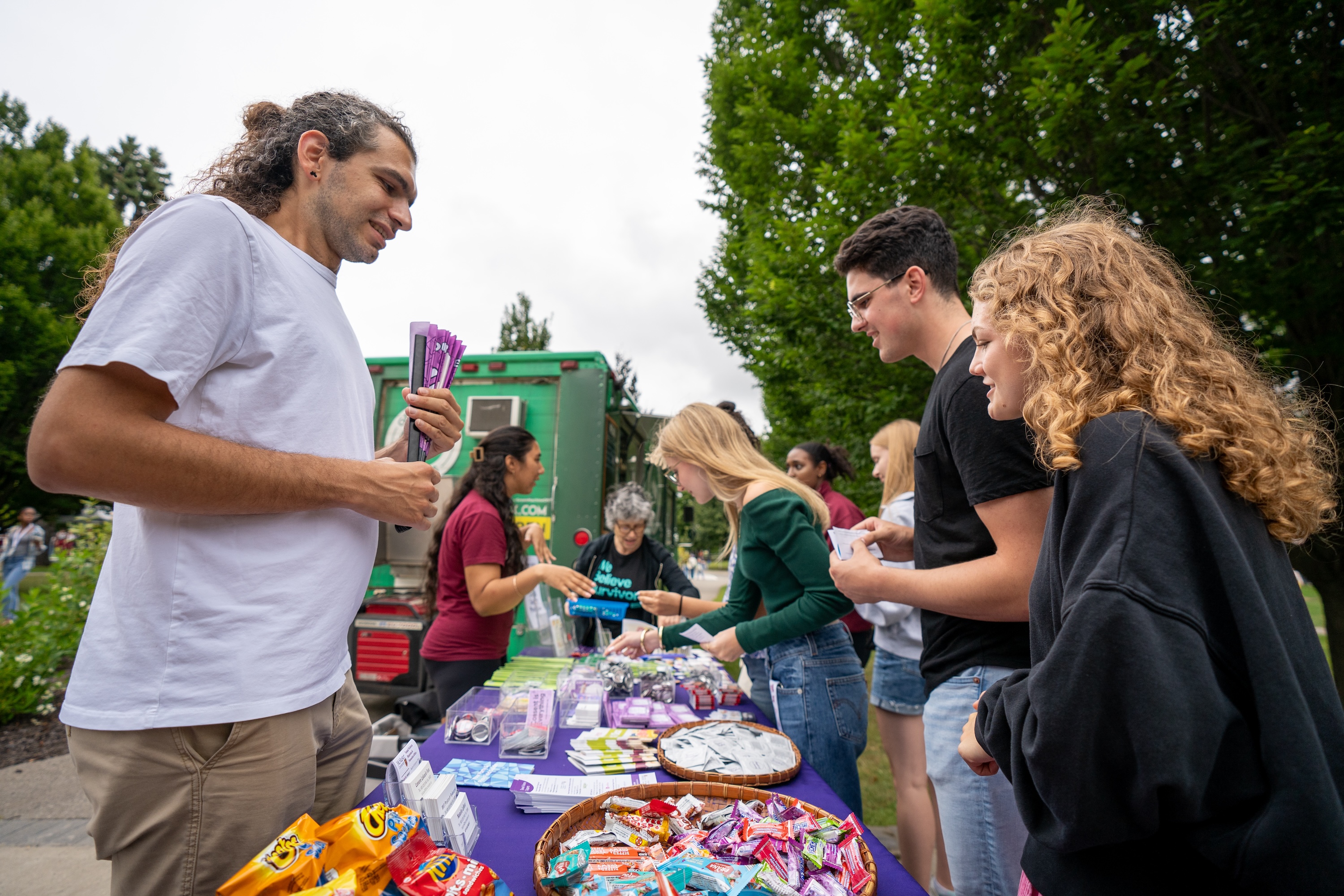 People stand on both sides of a table full of stickers and other swag outdoors on campus.