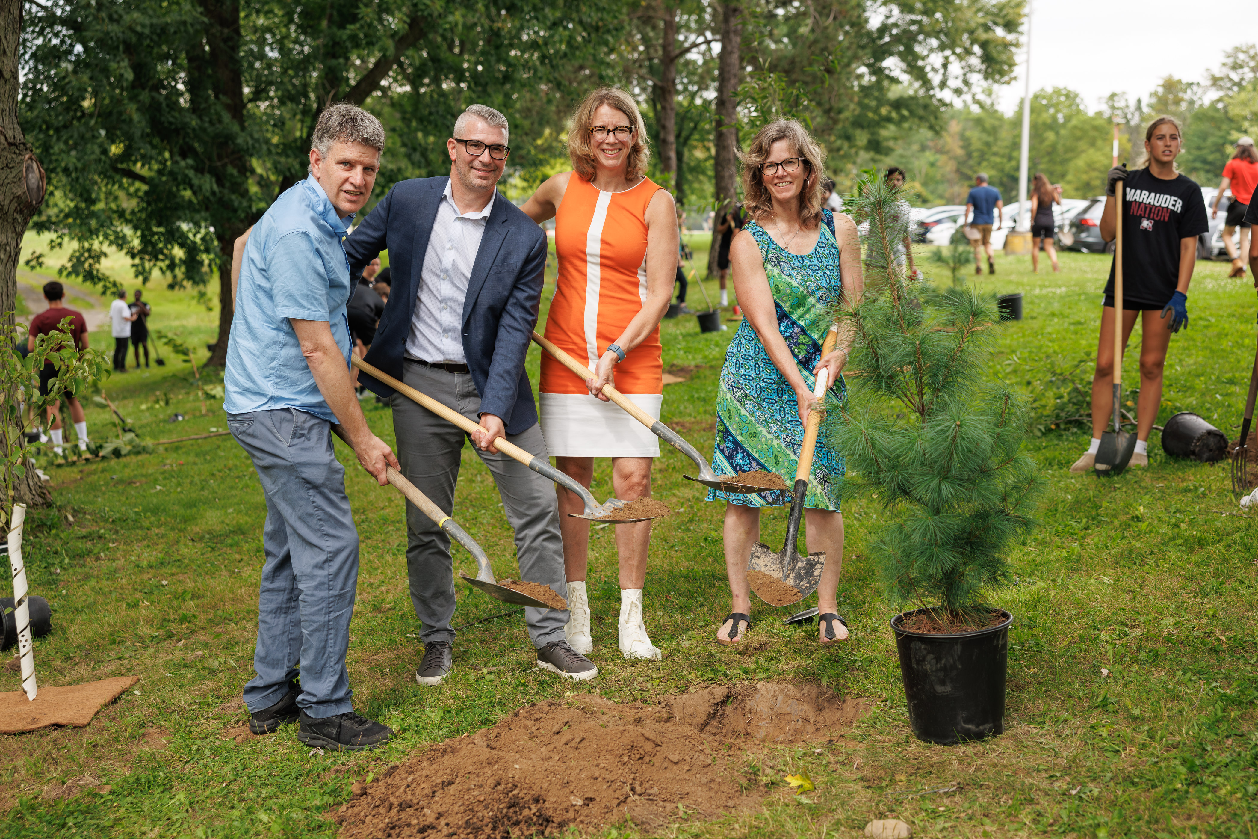 Four people holding shovels in front of a hole in the ground. There is a potted tree beside the hole.