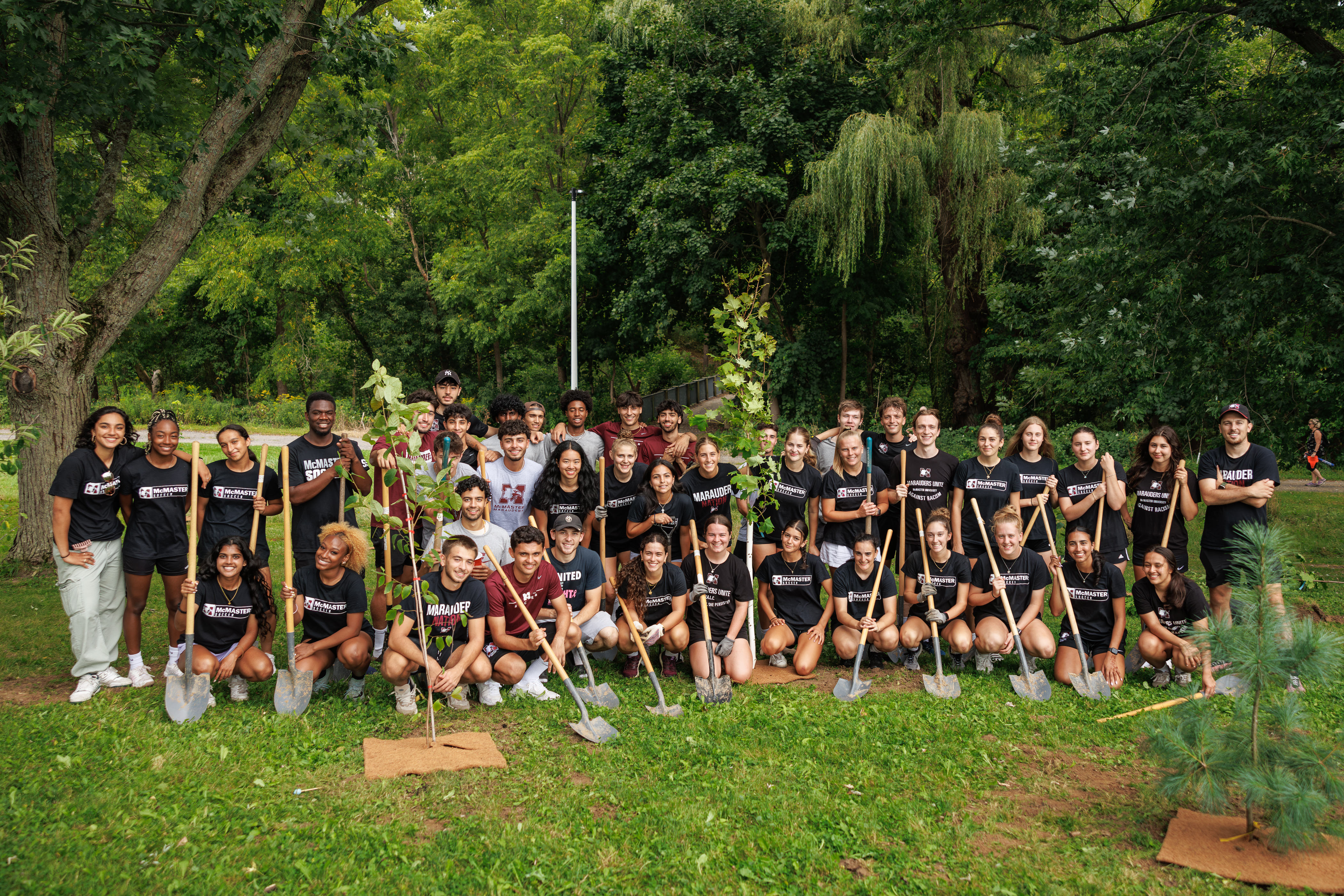 A large crowd of McMaster students in McMaster-branded clothing posing for a photo outdoors. Many are holding shovels.