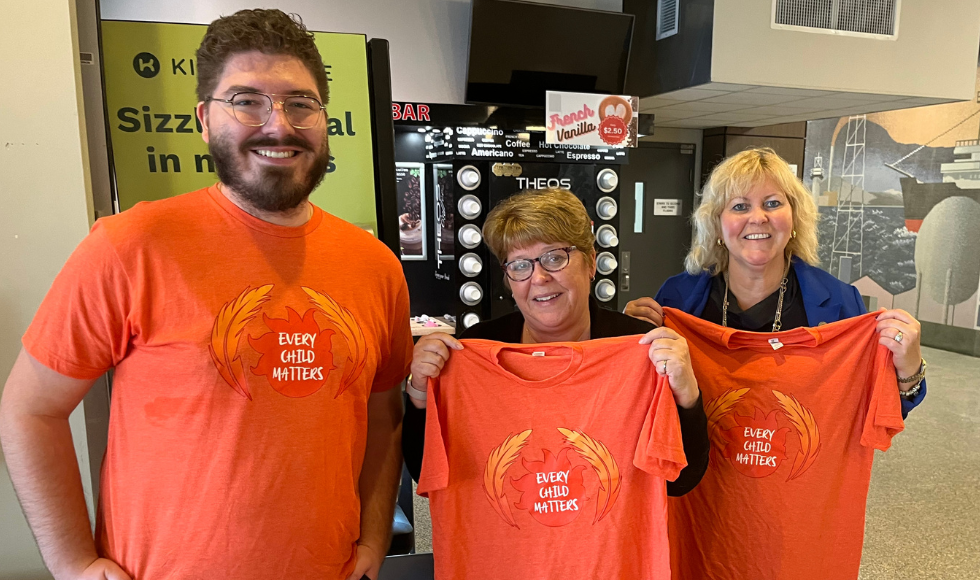 3 smiling people wearing orange Every Child Matters shirts in the lobby of the Hatch building on campus.