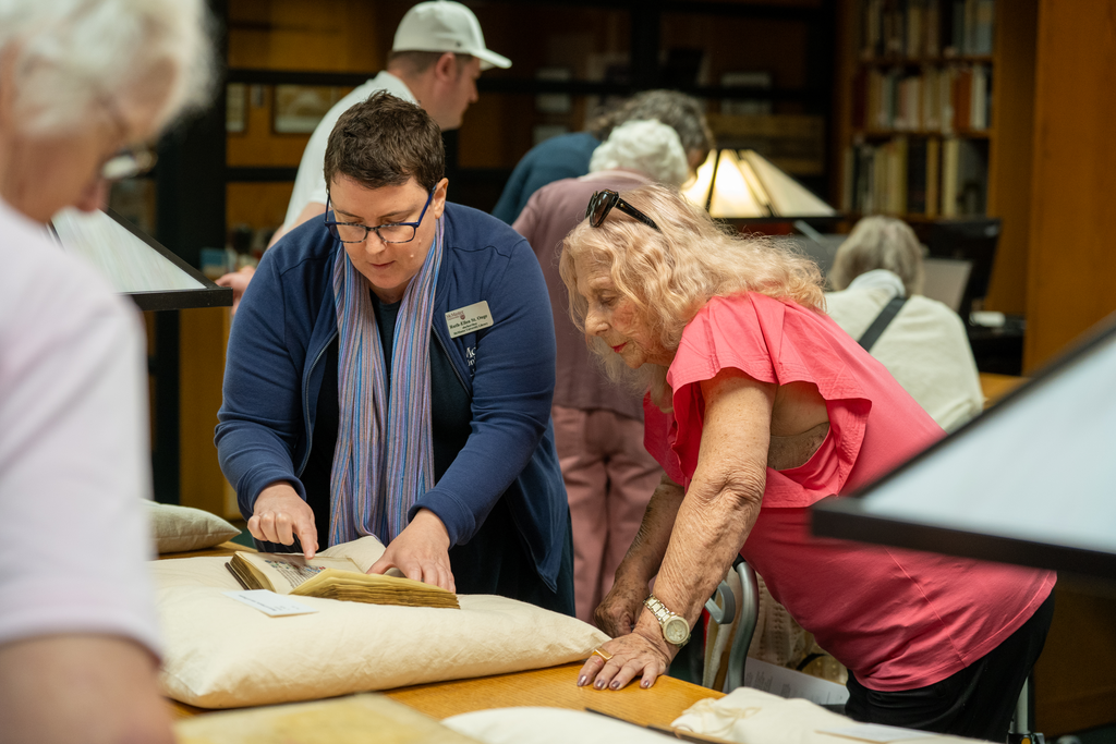 Two people at a table looking at a book with yellowed pages that’s sitting on top of a pillow 