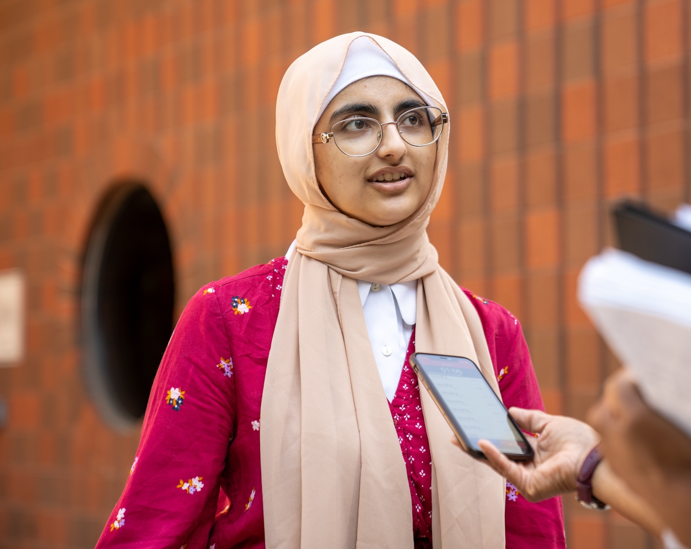 A woman wearing a hijab and a red and white top talks. A hand is holding a smartphone in front of her to record her voice.