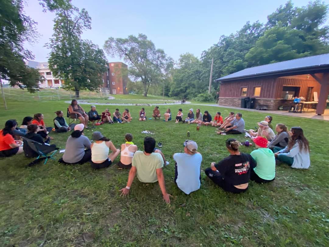 A large group of people sitting in a circle on the ground. One person in the circle is seated on a chair. 