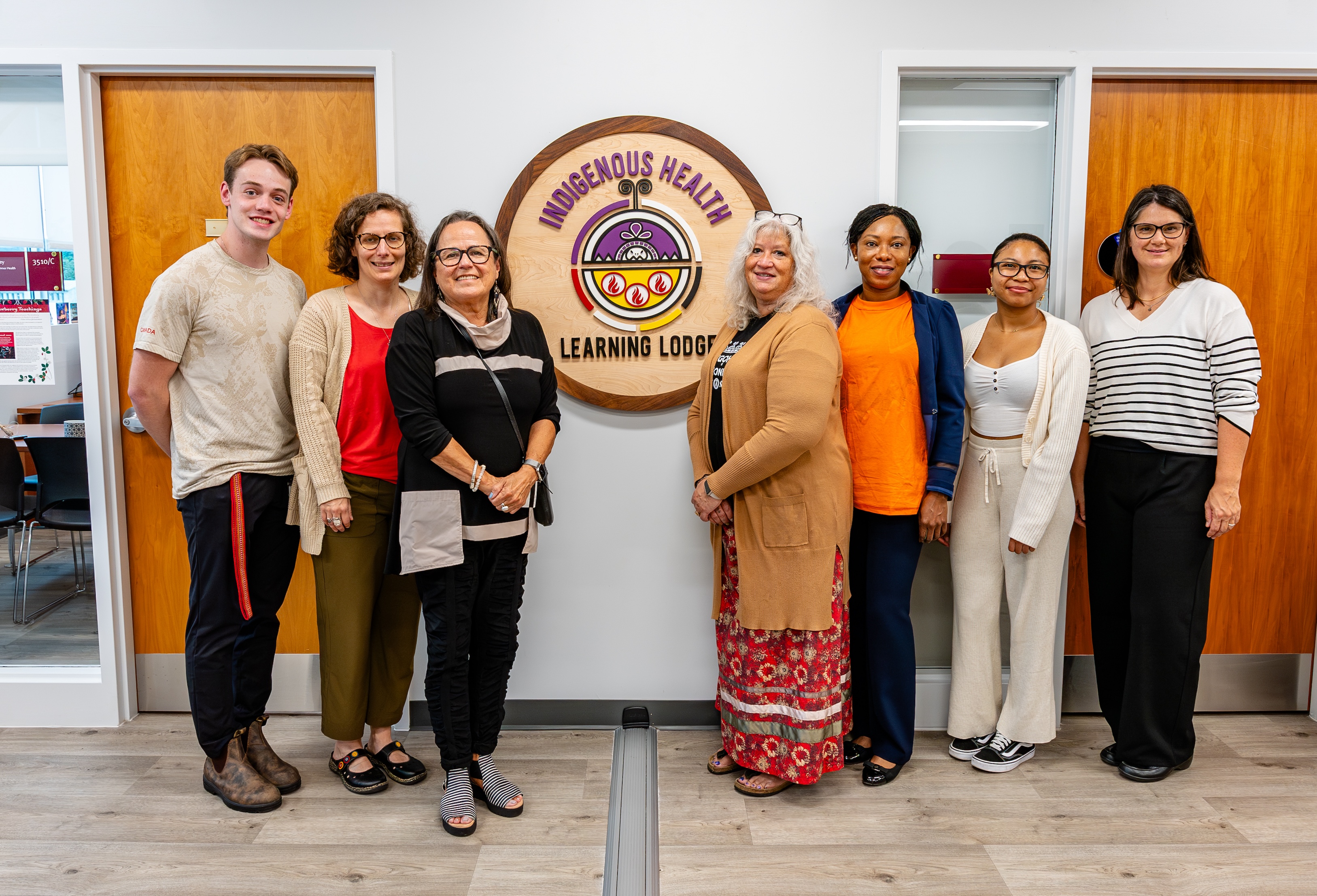 A group of smiling people flanks the Indigenous Health Learning Lodge emblem on the wall in the hallway.