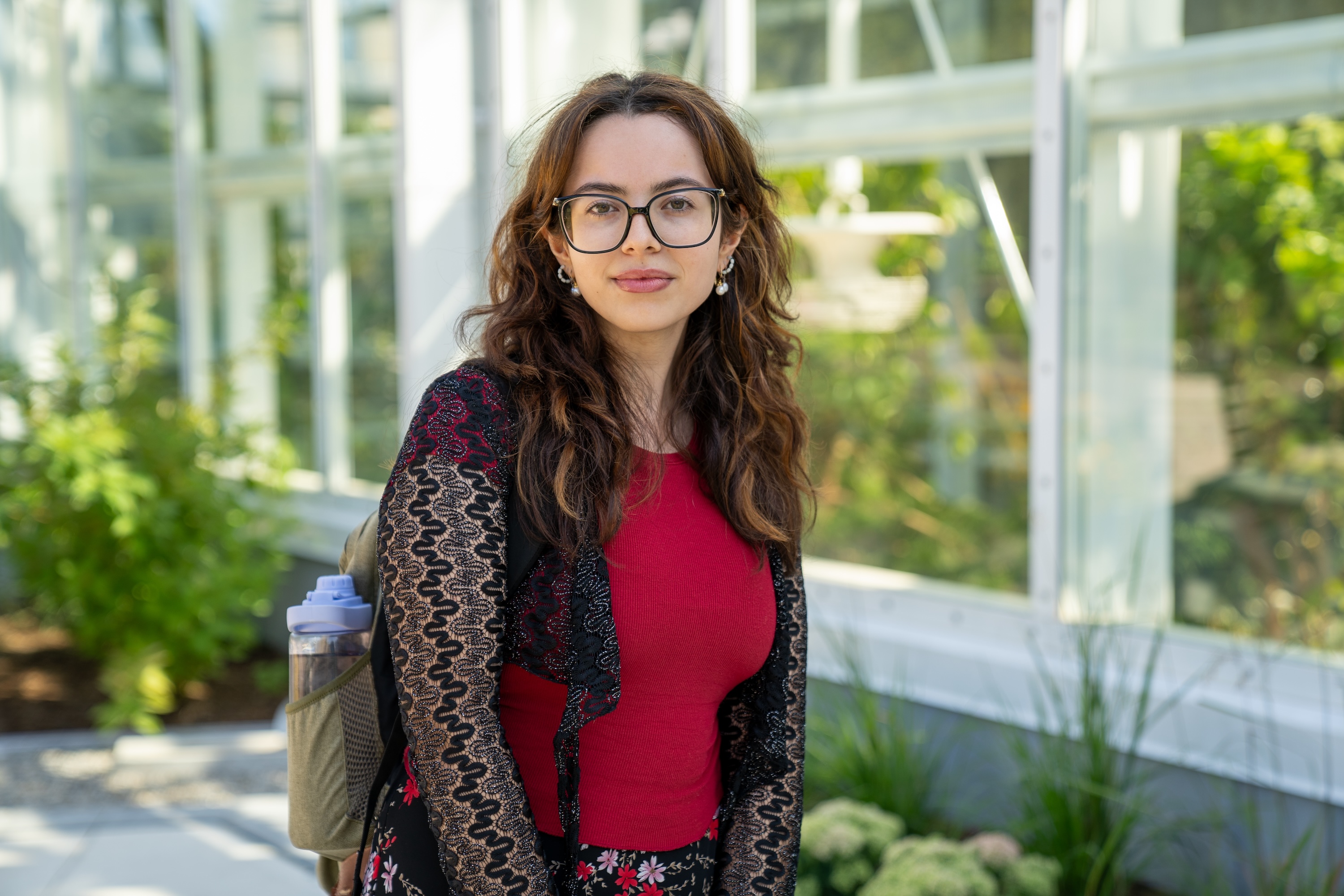 A person with long dark hair, glasses and a backpack wearing a red top with a black shrug over it, standing in front of the greenhouse building.