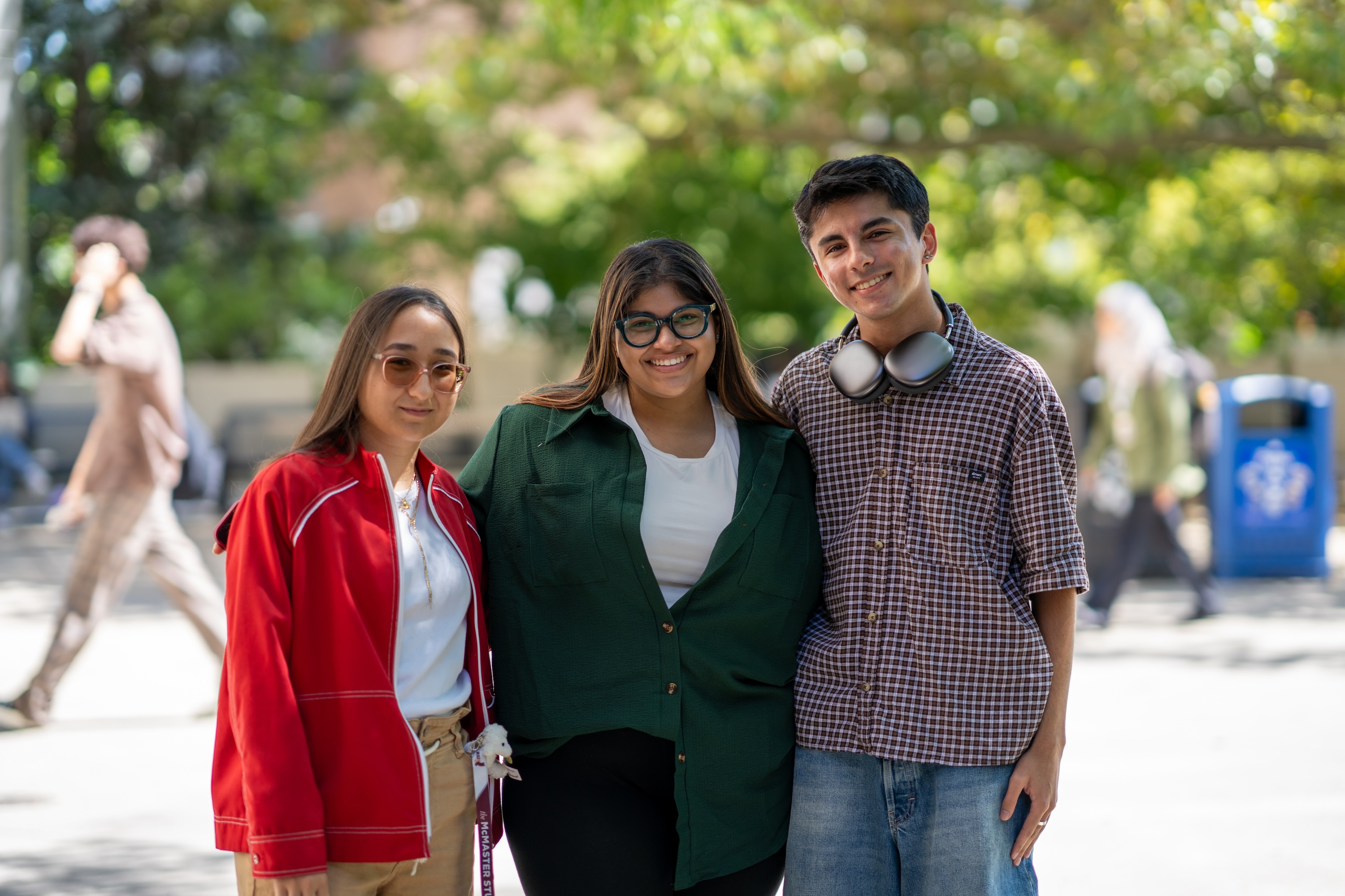 Three smiling students standing with their arms around each other's shoulders or waists in front of greenery in the Arts Quad on campus. 