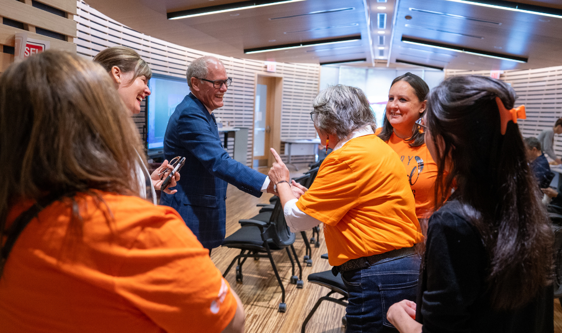 A man in blue and a woman in an orange T-shirt smile and shake hands in a group of 5 smiling people in the Ceremonial Room on campus.