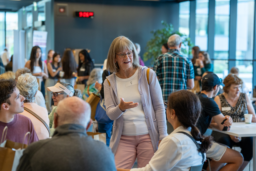 A woman standing in a room full of people while engaged in conversation with a student seated at a table 