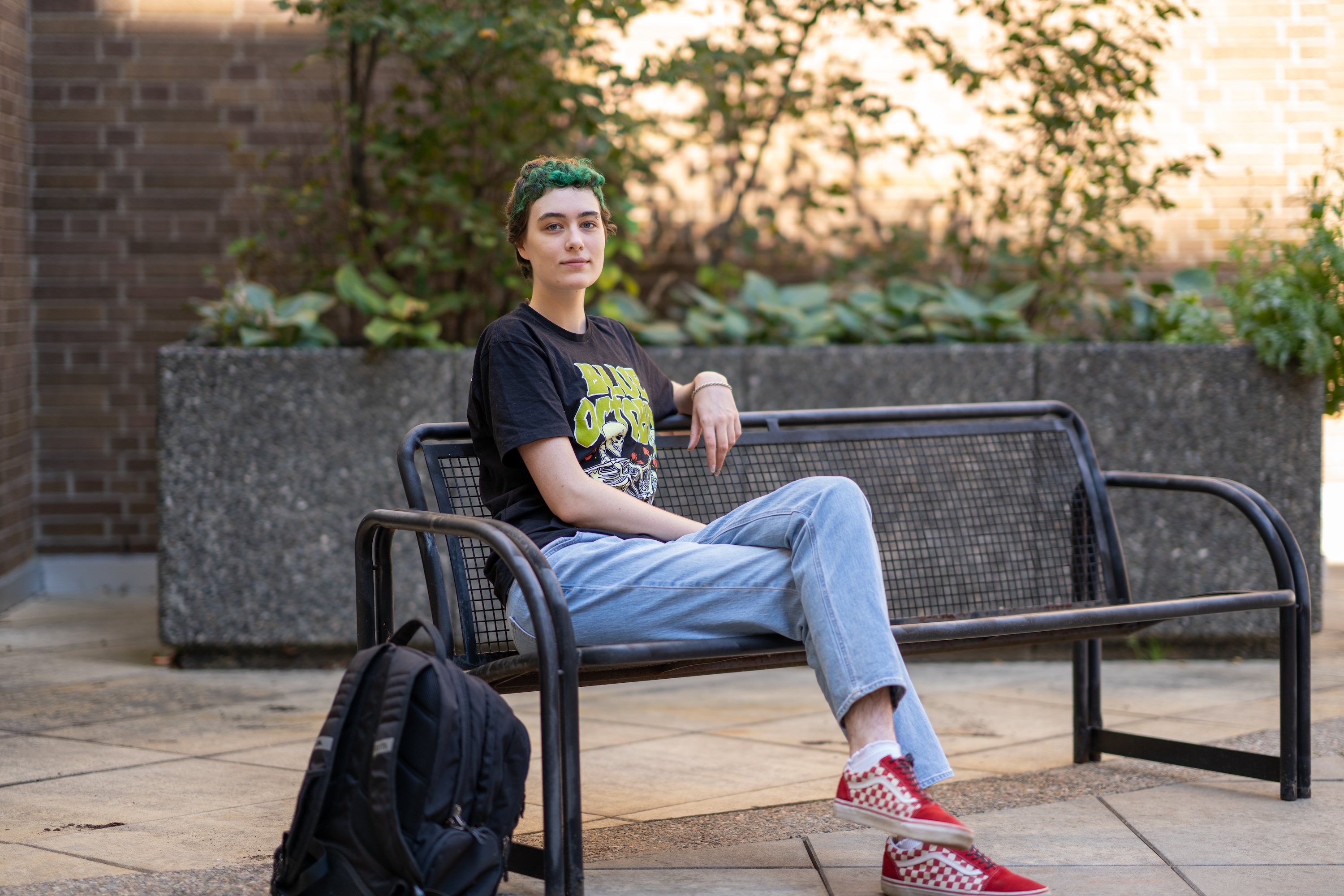 A student with short hair in a dark Tshirt, blue jeans and red sneakers sits on a bench on campus, their backpack leaning against the bench.