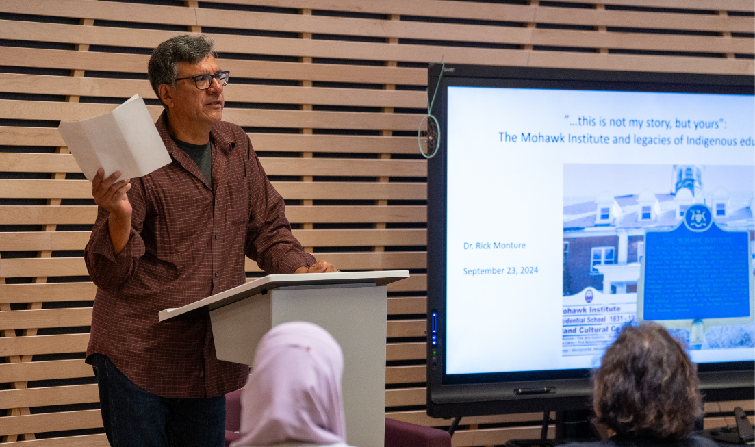 Rick Monture holding up paper and speaking at a lecturn in the Ceremonial Room, beside the screen showing the Truth and Reconciliation presentation.