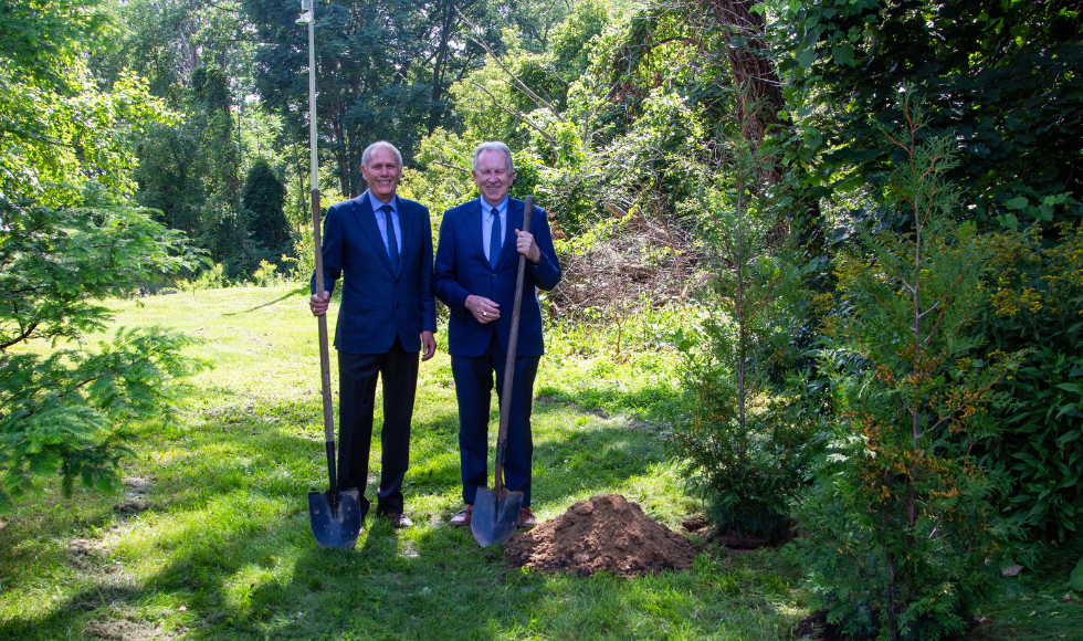 Two men in blue suits holding shovels and smiling at the camera while standing beside a small hole in the ground. 