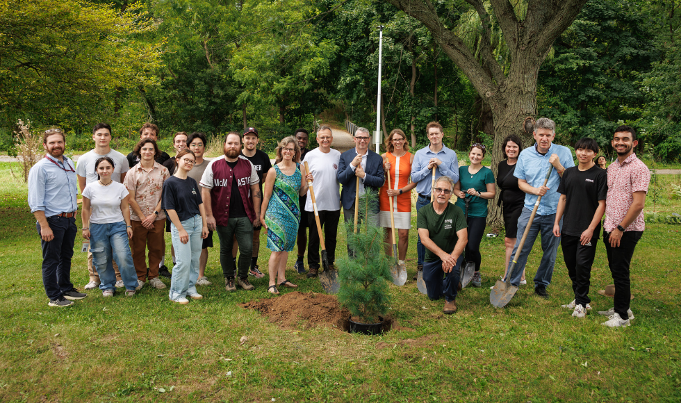 22 people smiling at the camera while standing outdoors in front of a potted tree that has been placed in a hole in the ground.