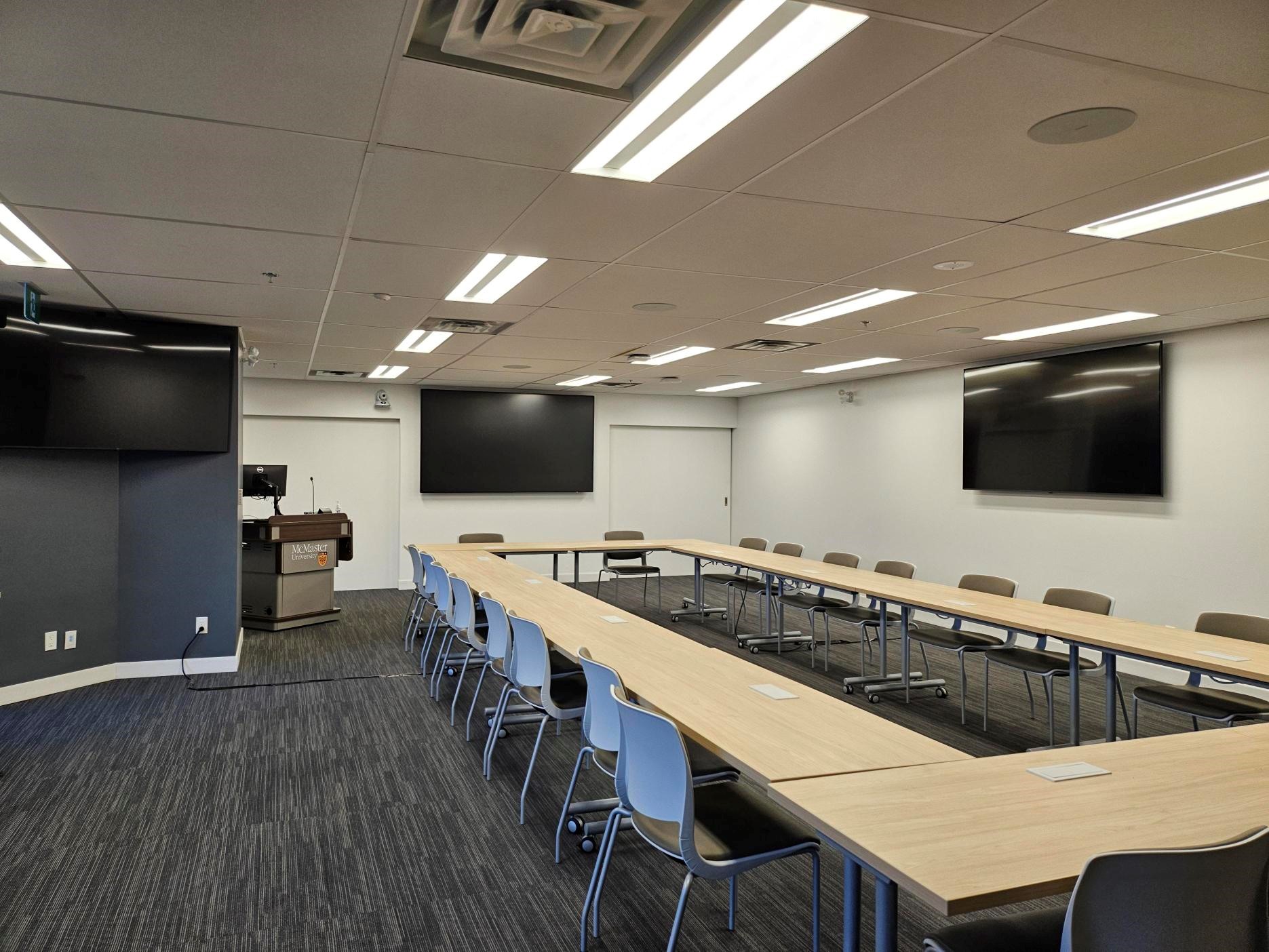 A boardroom with screens on the walls. There are a series of desks and chairs arranged in a rectangular format in the centre of the room. 