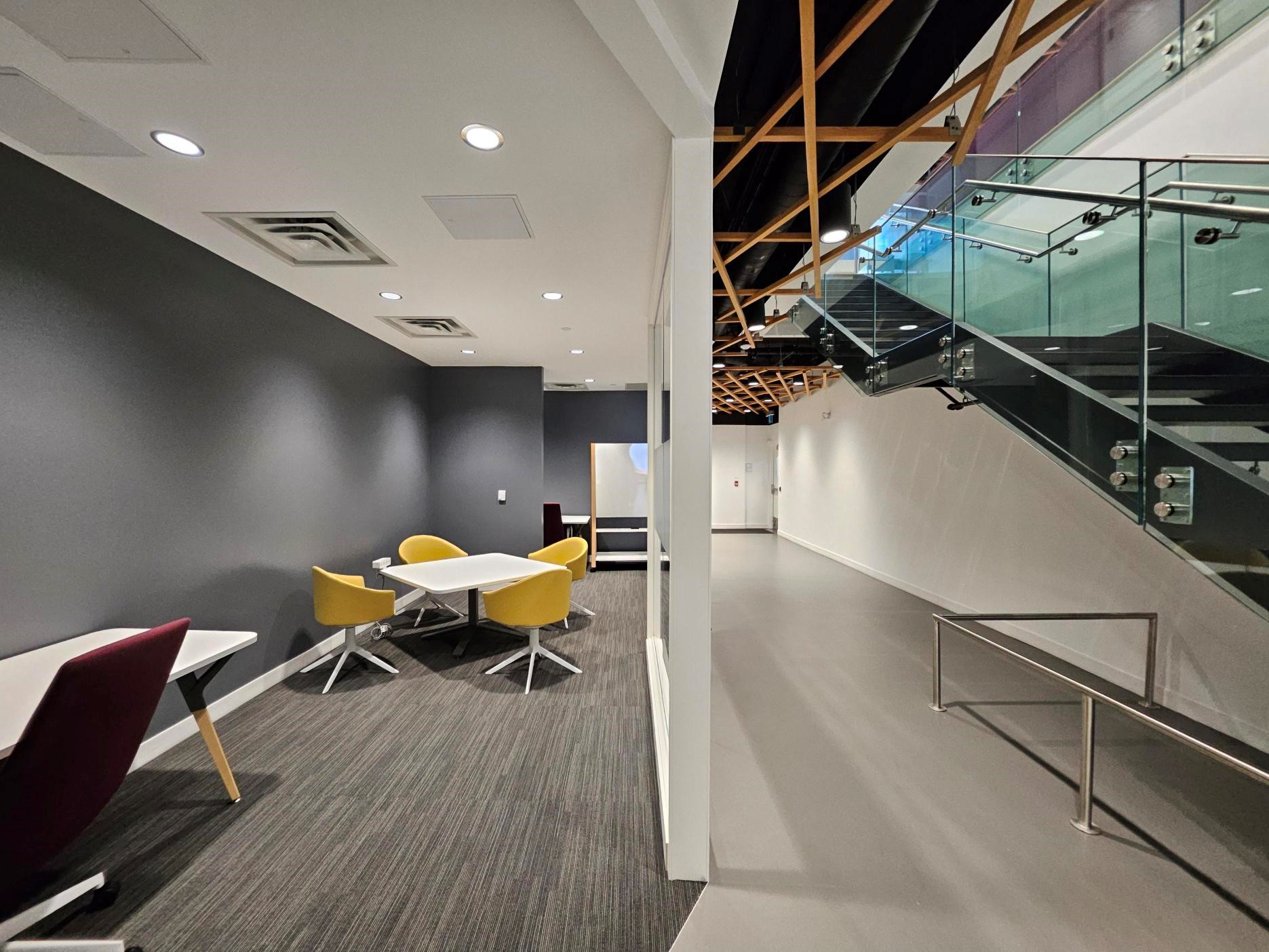 A section of a building with a modern glass staircase, and a lounge area with a whiteboard and different work areas. 