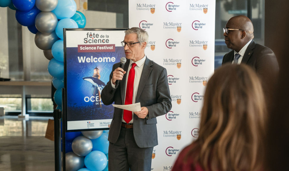 Bertrand Pous speaks into the microphone as Bonnie Ibhavo looks on. Behind them are signs advertising the Science Festival and the McMaster University logo. 