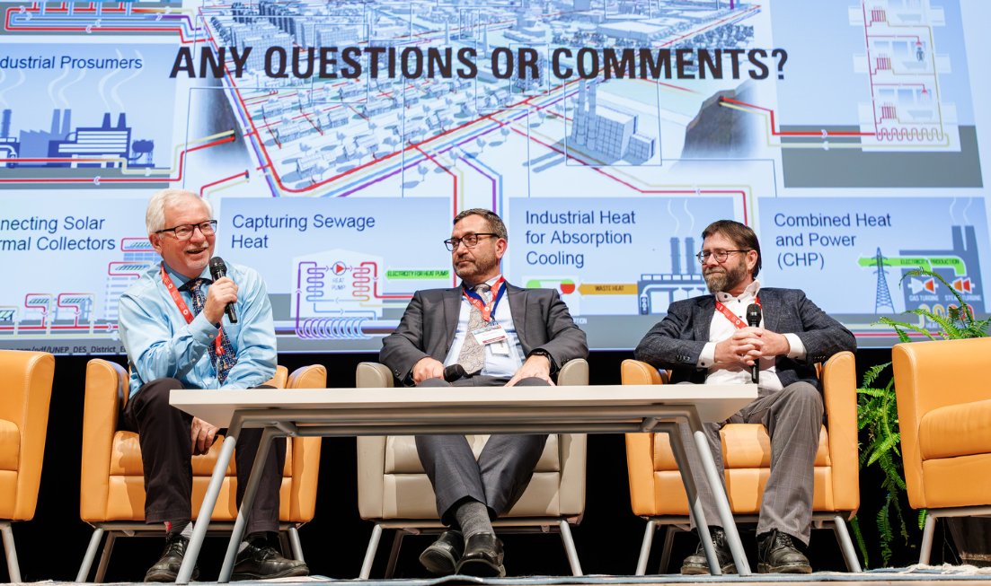 Three professors sit on stage for a panel discussion, in front of a massive screen showing their slide presentation.