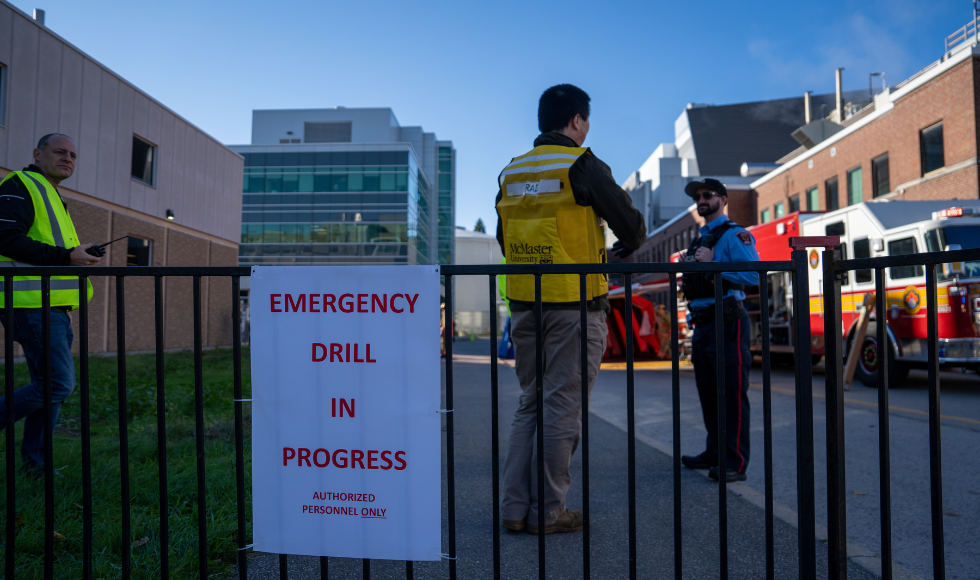 A sign fixed to a fence that reads, 'Emergency drill in progress. Authorized personnel only.' Behind the fence are three people and a firetruck.