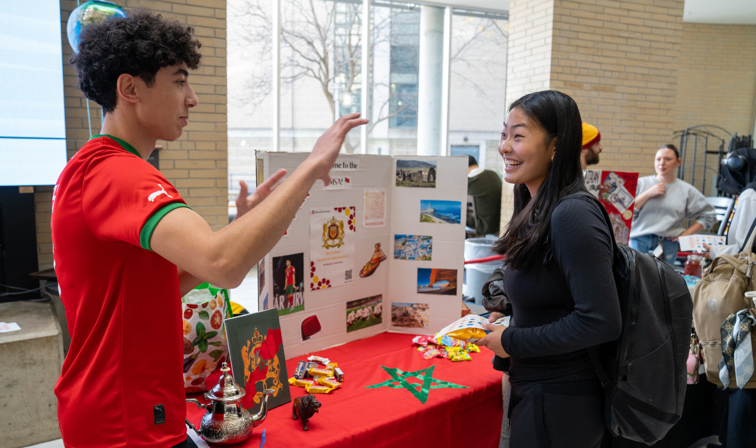 A student in a soccer jersey chats with a smiling student as they stand at a colourful table for the Moroccan Students Association in MuSC