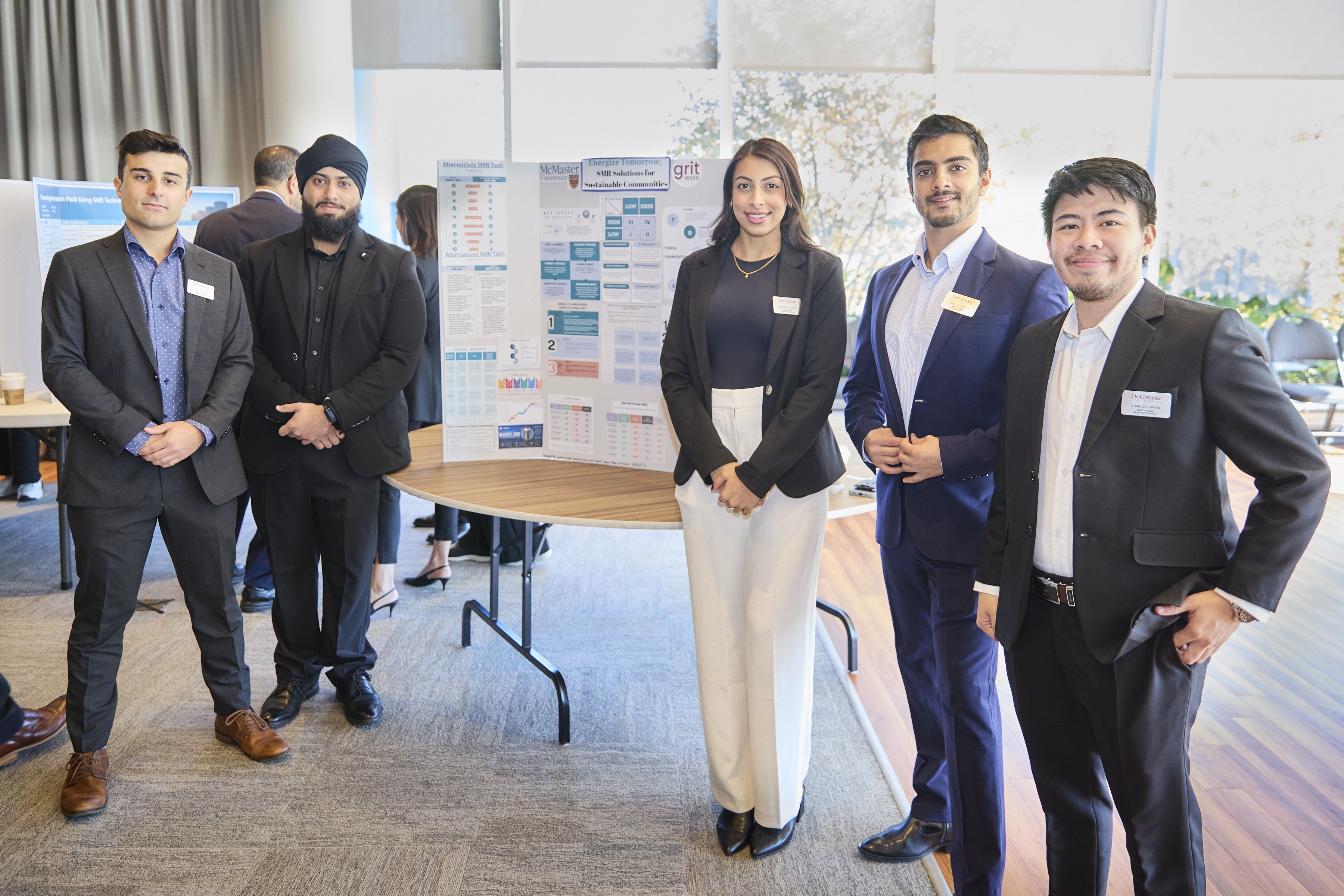 Five students in business suits stand beside a poster presentation.