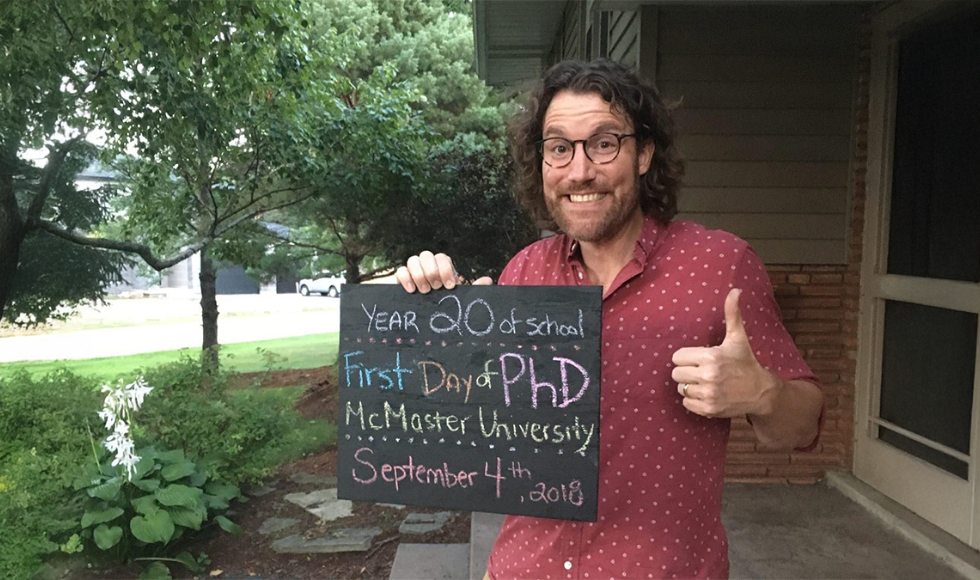 Aric Rankin giving a thumb’s up while holding up a chalkboard with text on it that reads, ‘Year 20 of school - First day of PhD - McMaster University - September 4th, 2018.’