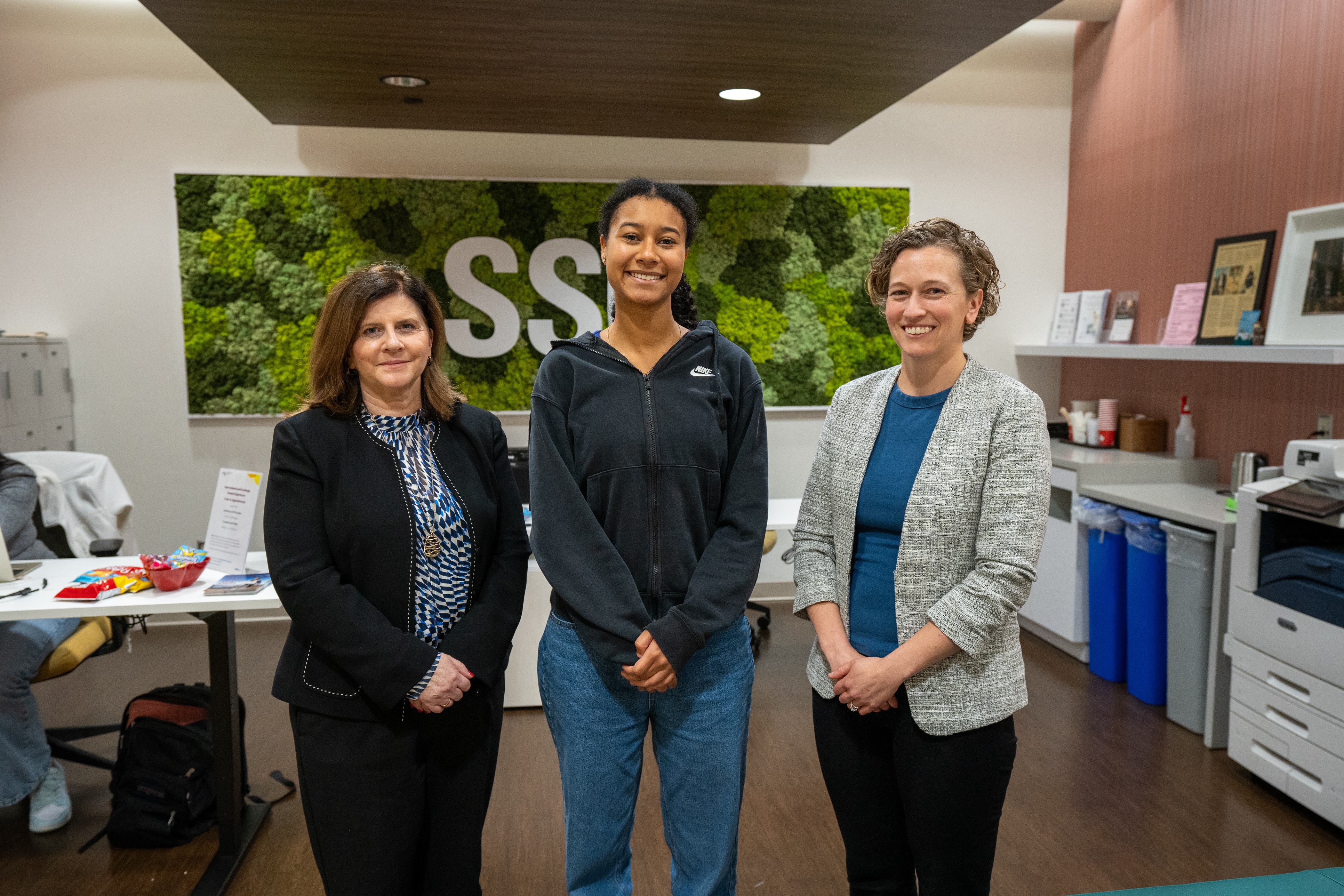 Three smiling people stand indoors in front of a large green wall with the letters SSC spelled out in white.