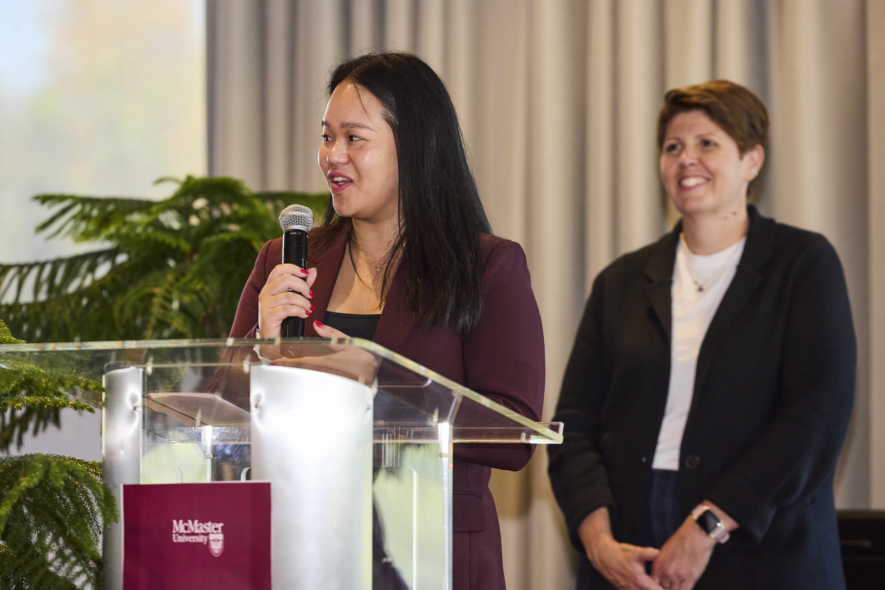 A person with long dark hair speaks into a mic at a McMaster-branded lectern, while a second person, in a suit, standing behind, smiles widely.