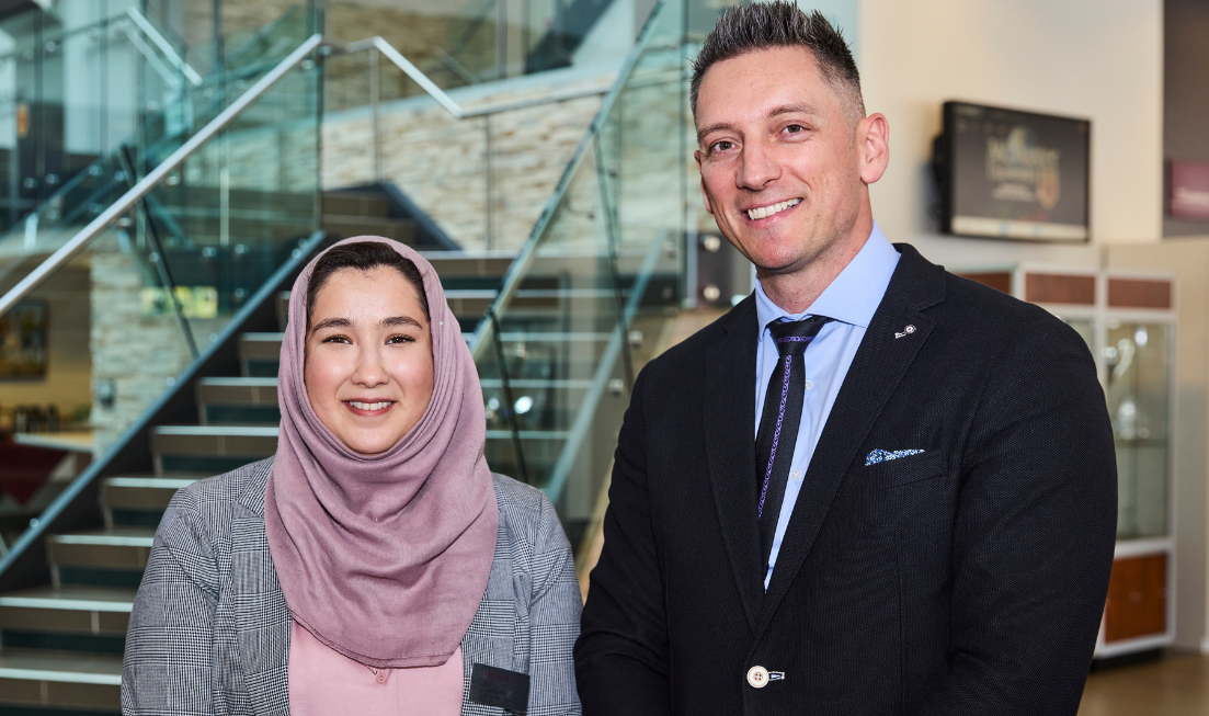 A woman in a hijab and blazer and a man in a suit smile for the camera in the lobby of a building.