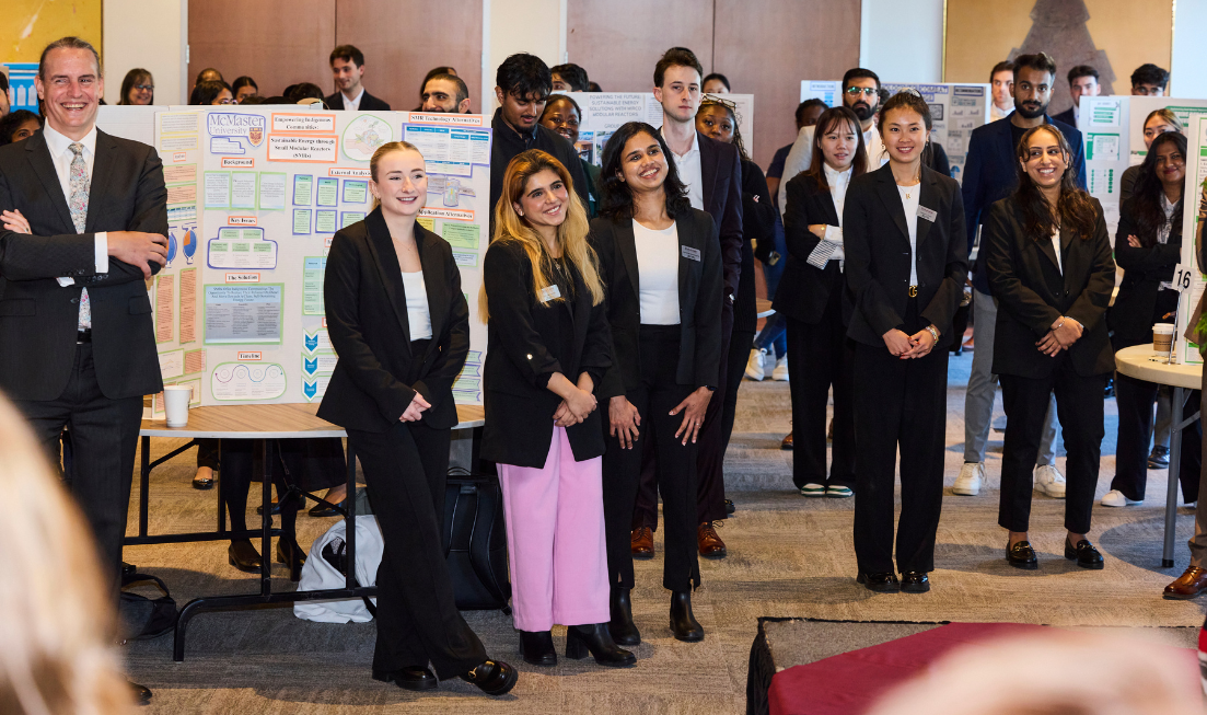 a large group of students in business suits standing in front of posters and whiteboards watching something.