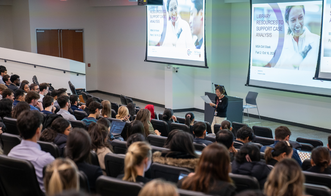 A speaker addresses a theatre-style classroom with a slide show on the big screens hanging right behind them on either side.