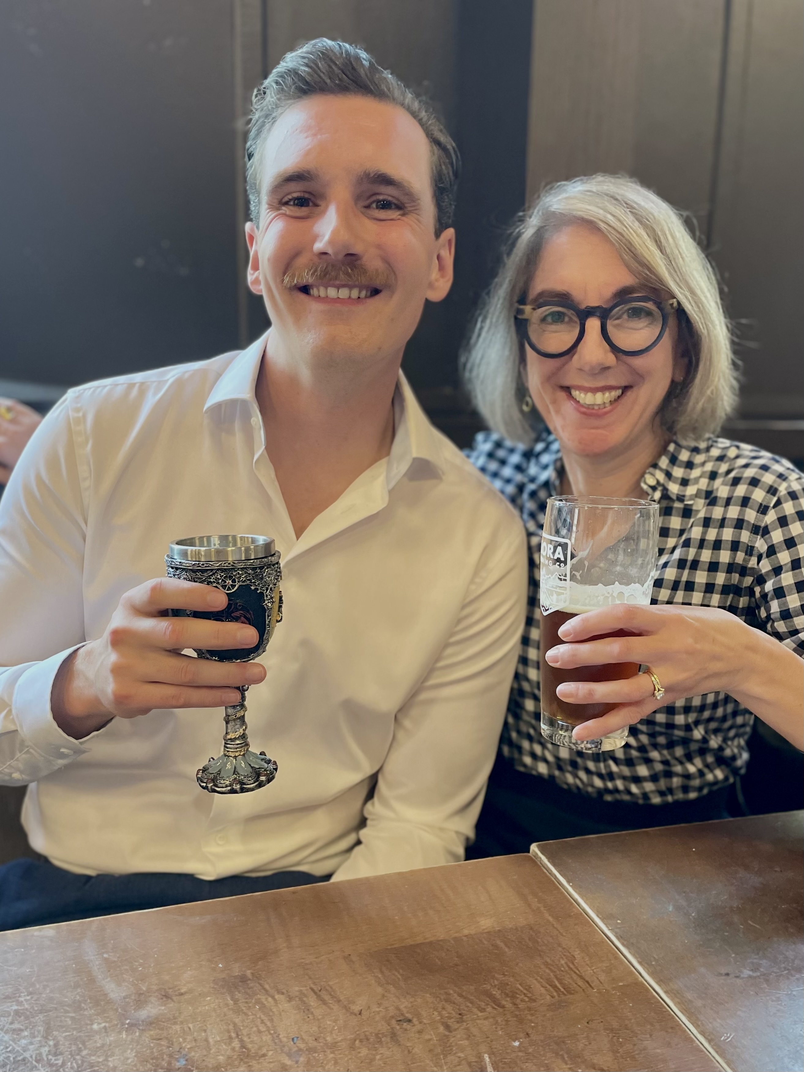 Ryan Heyden seated beside Dean of Humanities Pamela Swett. Both are smiling directly at the camera. Heyden is holding a chalice, while Swett is holding a pint of beer. 