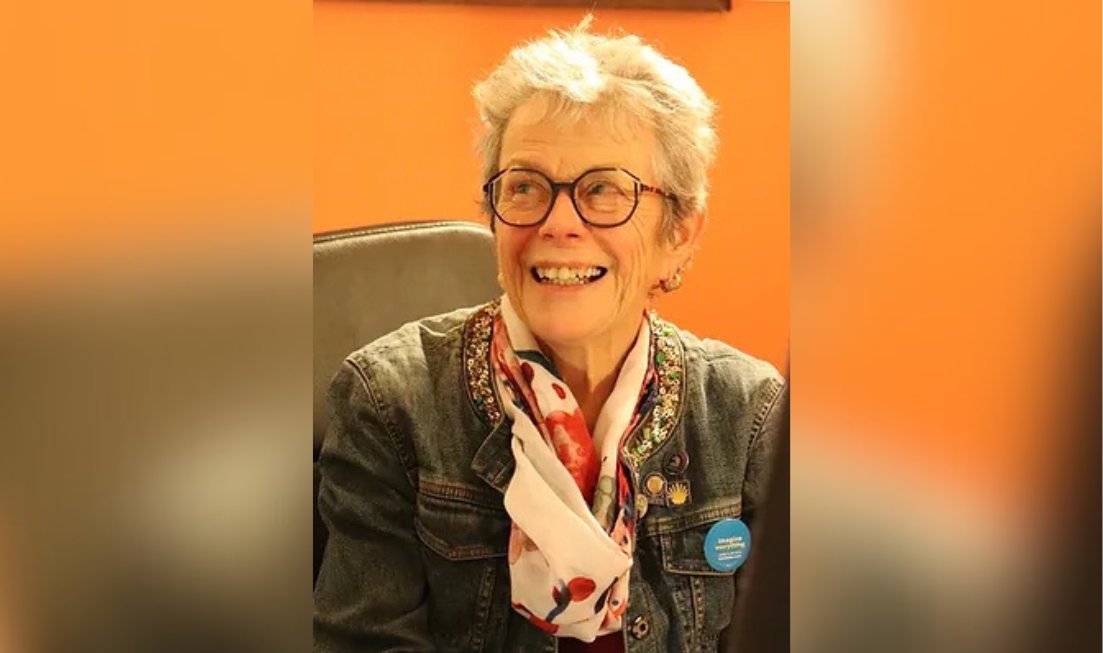 Jean Marmoreo, wearing glasses, a denim jacket and a red and white scarf, looks off to the side with a wide smile.