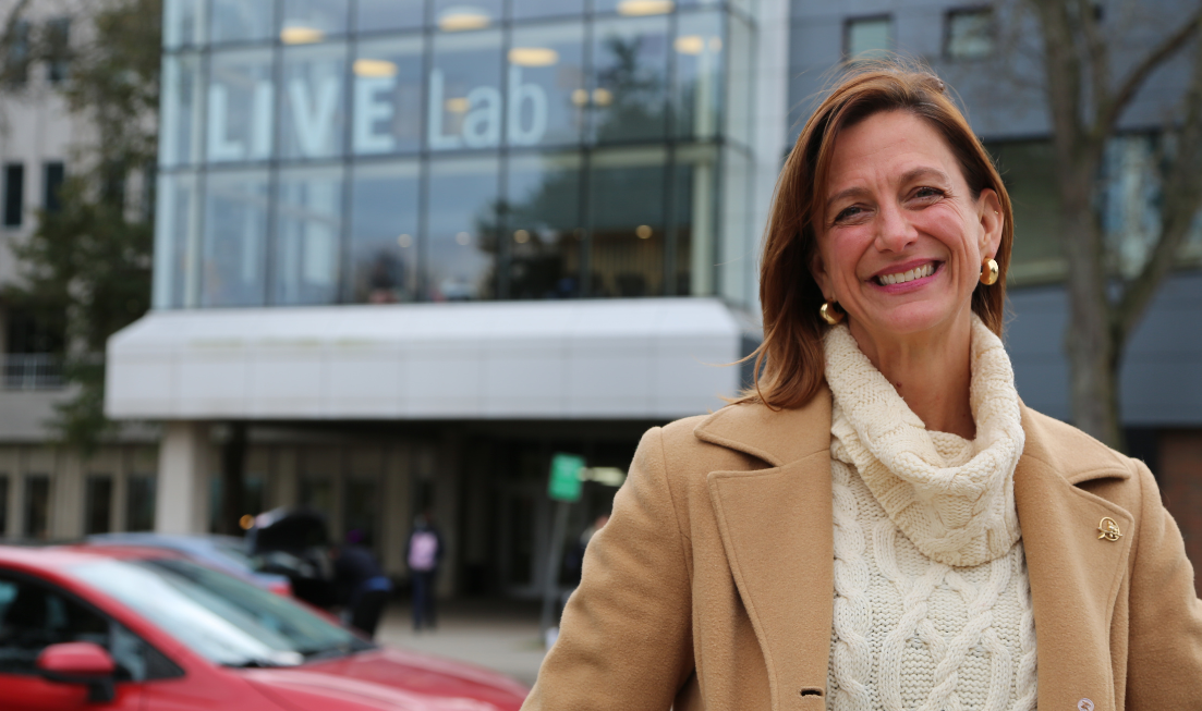 Smiling Rachael Finnerty stands in the foreground in a turtleneck sweater and a tan coat, with the LIVE Lab's building with its distinctive labelled windows in the background.