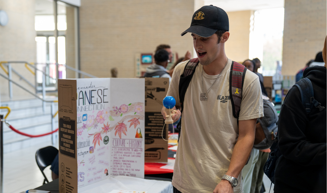 A young person in a ball cap and T shirt, wearing a backpack, plays with a japanese Kendama beside a table with a "Japanese Student Club" sign on it. 