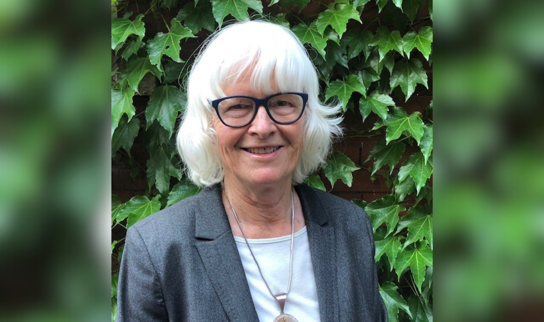 Head and shoulders of a smiling white-haired person with glasses, wearing a white top under a grey blazer, with a chunky pendant on a chain, standing against a leafy green backdrop.