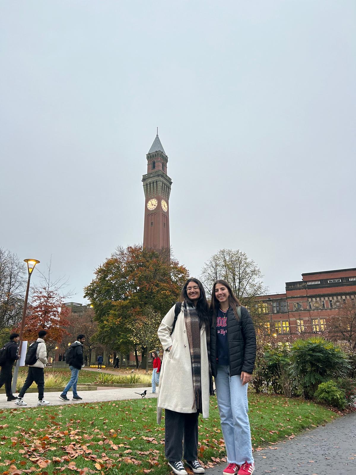 Two smiling students in casual clothes and jackets stand outdoors during the fall in front of a university building and a clock tower.