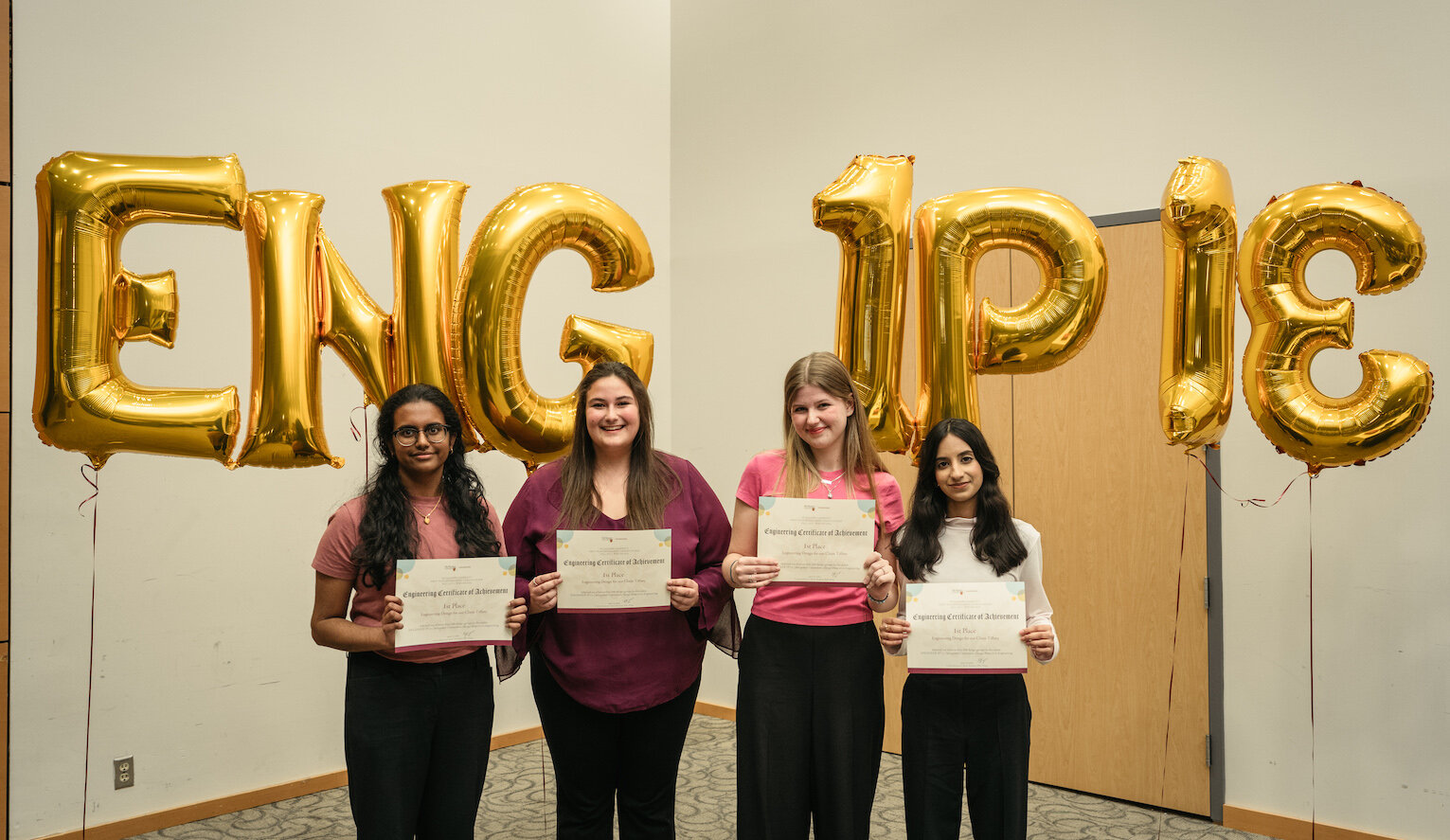 Four people standing and holding up certificates. Behind them are large gold balloons that spell out ENG 1P13.