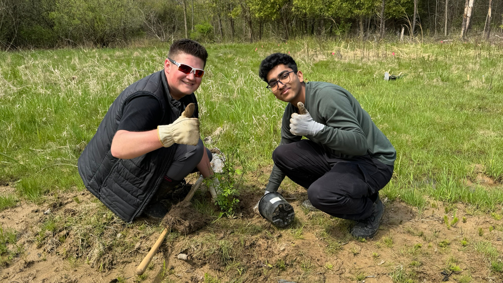 Two students crouched down planting a tree. They are both turned towards the camera, giving a thumbs up. 