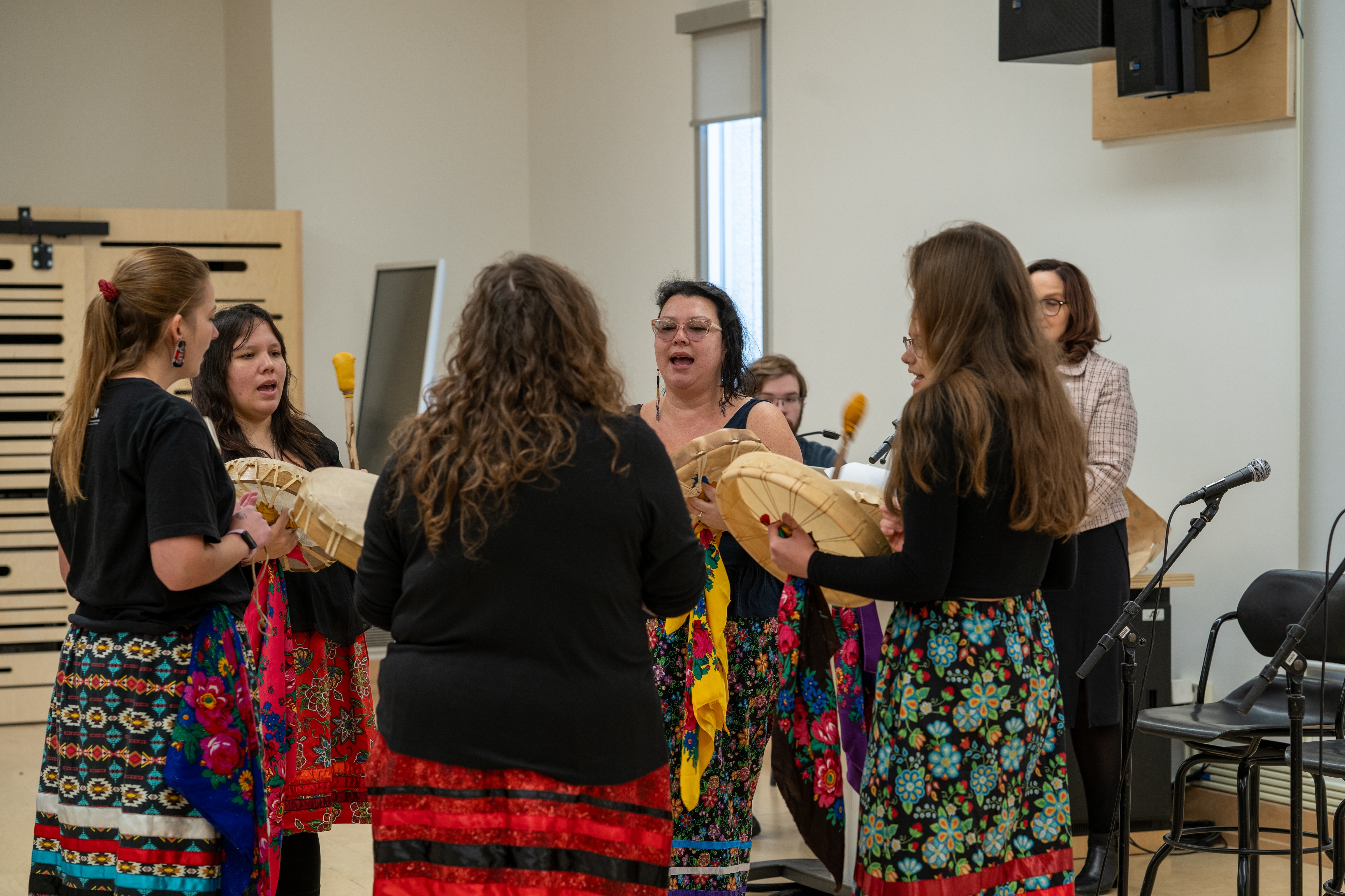 Drummers from the student-led Kindred Spirits Drum Circle, in ribbon skirts, stand in a circle playing Indigenous drums. 