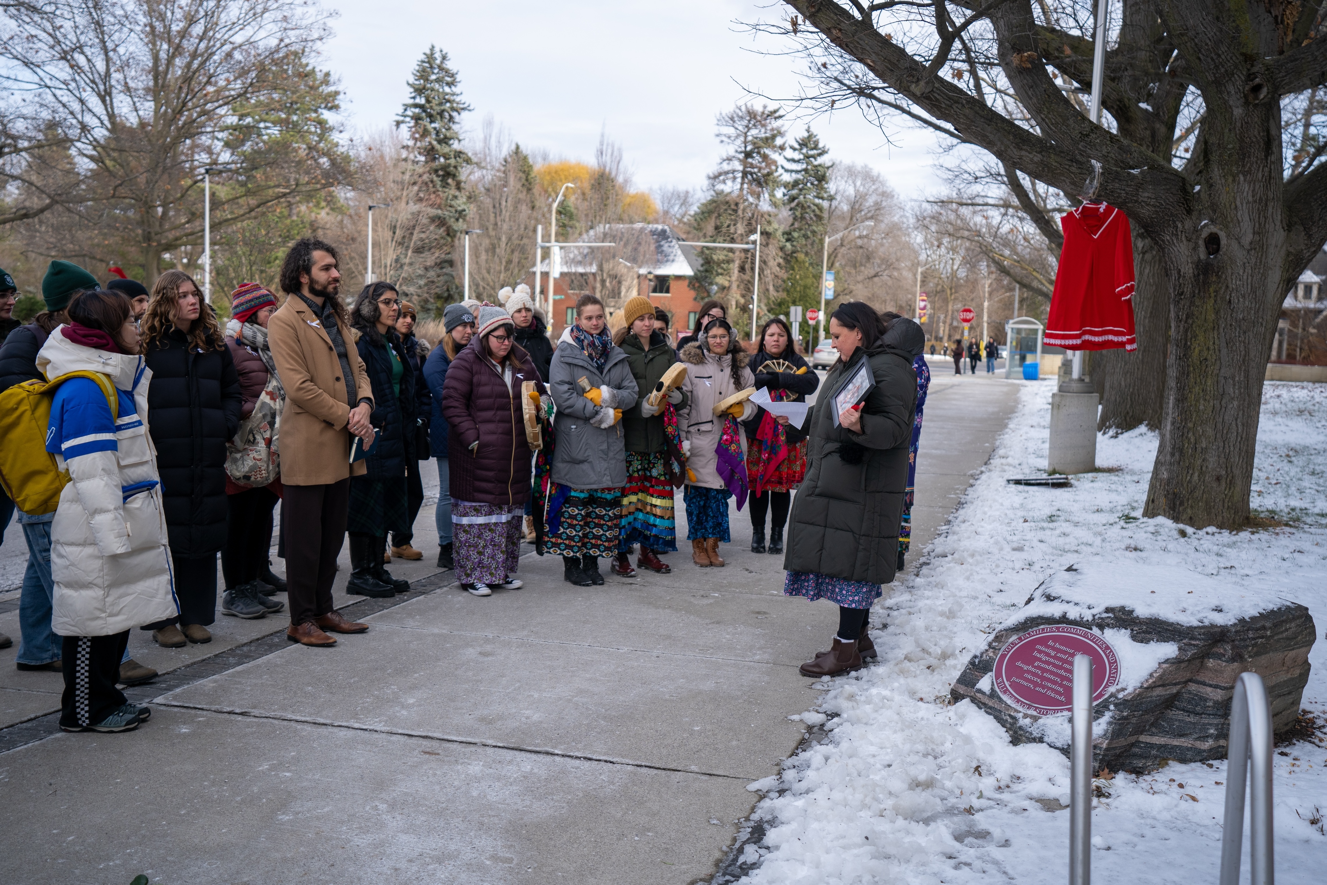 People stand in a semicircle at a memorial stone outside L R Wilson Hall, with a red dress hanging from a tree in the background. 