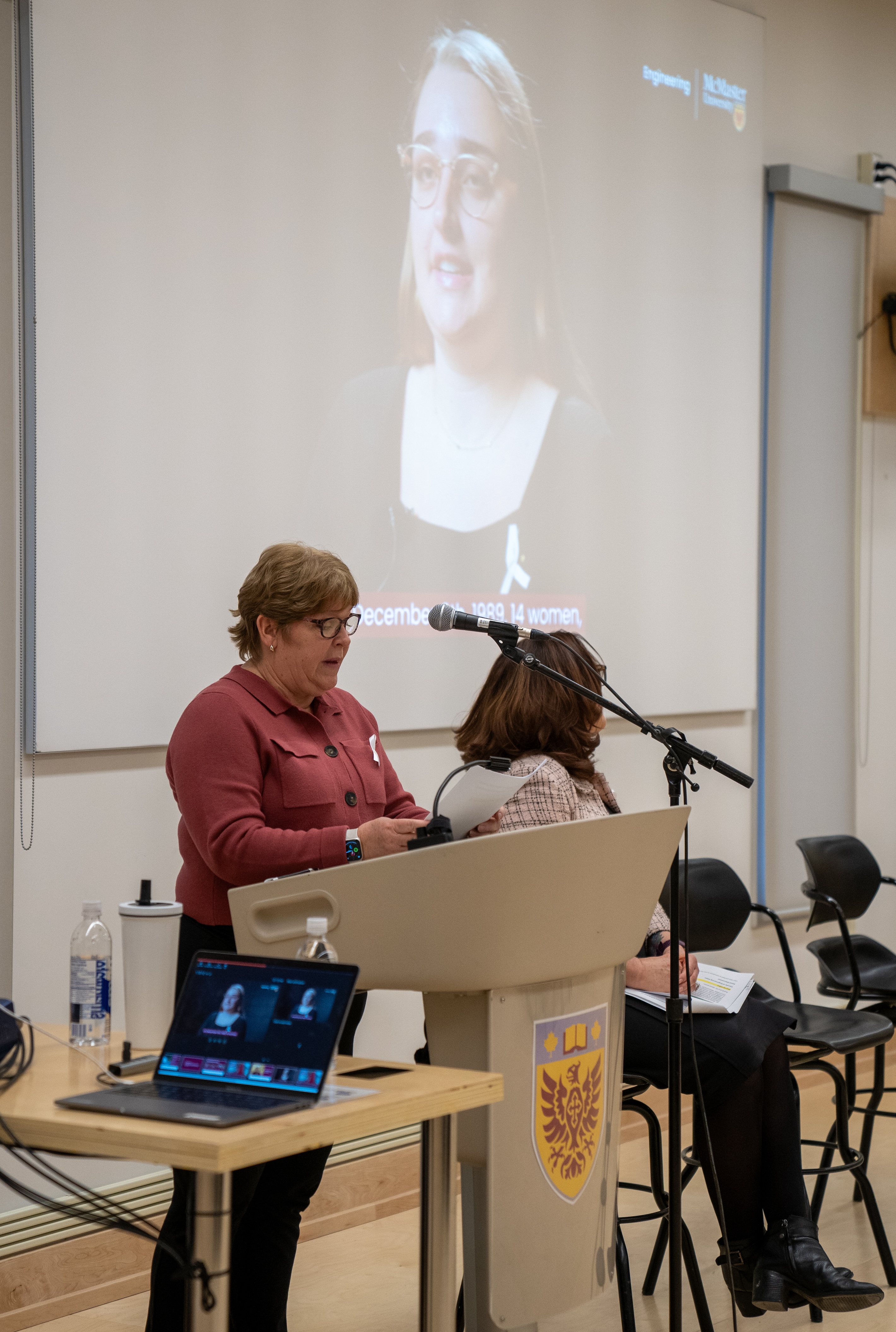 Engineering Dean Heather Sheardown speaks at a podium while a remembrance video plays on the screen behind her at a Dec. 6 event in a classroom.