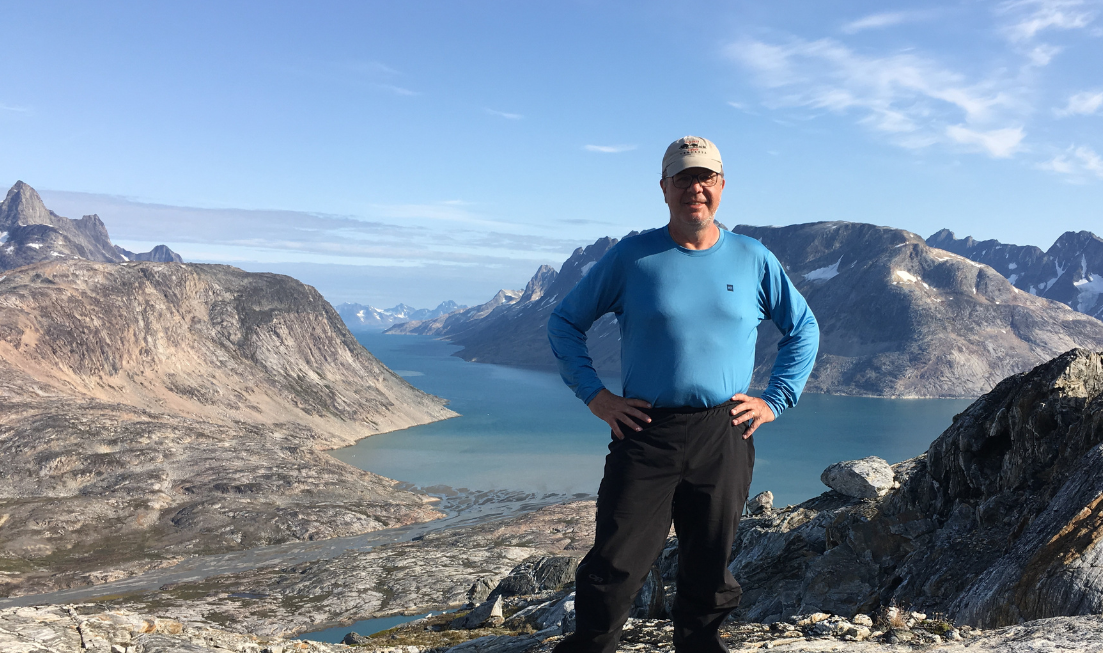 Full-length shot of Doug Welch standing on a very high rocky spot, with a lake or glacier or river behind him, framed by mountains.