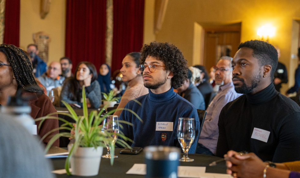 Many people are seated at tables throughout a large conference space. They all seem to be listening intently and watching something at the front of the room. 