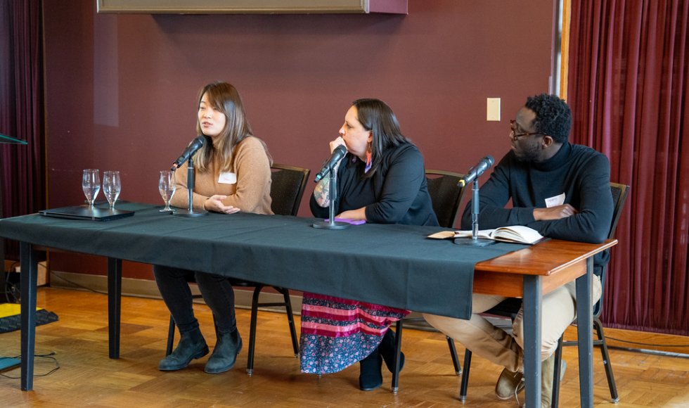 Three people seated at a table, each with a microphone in front of them and facing the same direction. The woman on the left is speaking. 