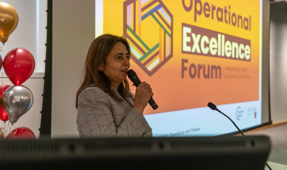 A woman in a blazer speaks into a microphone. She is standing at a podium in front of a presentation screen, which reads "Operational Excellence Forum." 