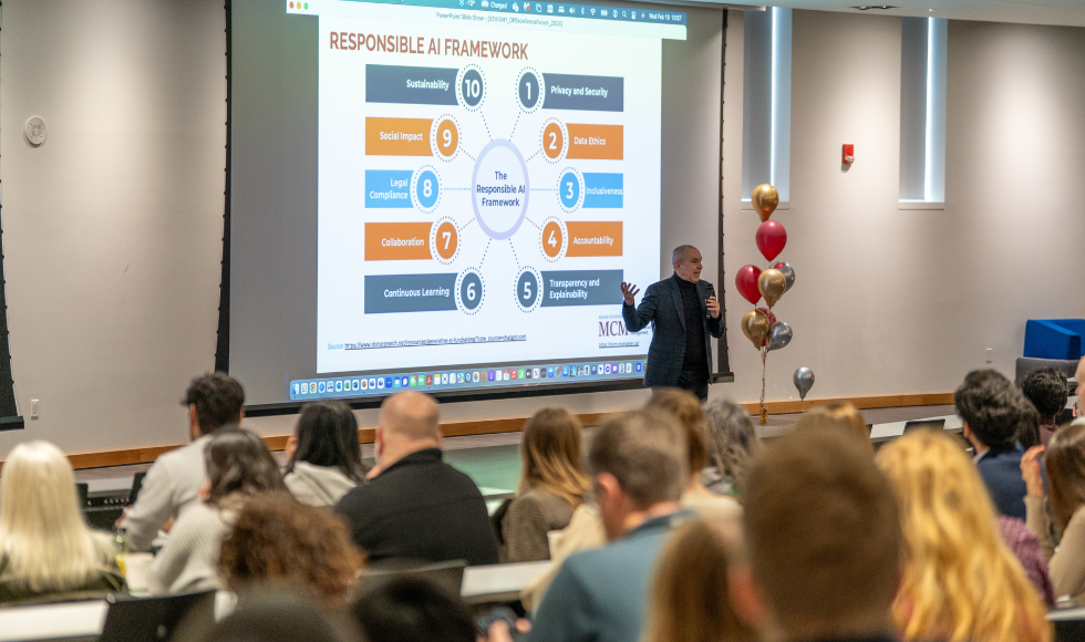 At the front of a crowded classroom, a man stands in front of a presentation screen, speaking into a microphone. 