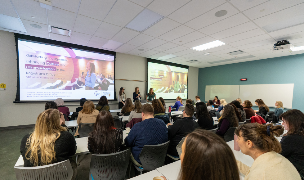 At the front of a full classroom, three women stand in between two large presentation screens. They are facing the classroom, presenting. 