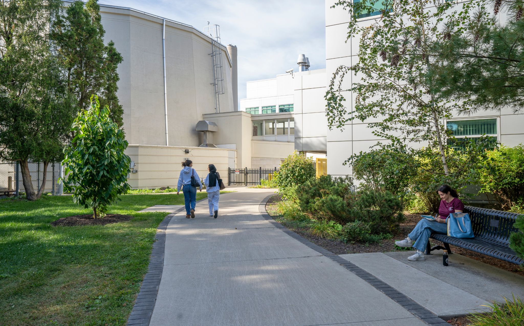 A wide concrete path runs through an outdoor space. A person sits on a bench on the right side of the path, and further away, two people walk down the path, heading towards a large concrete building. 