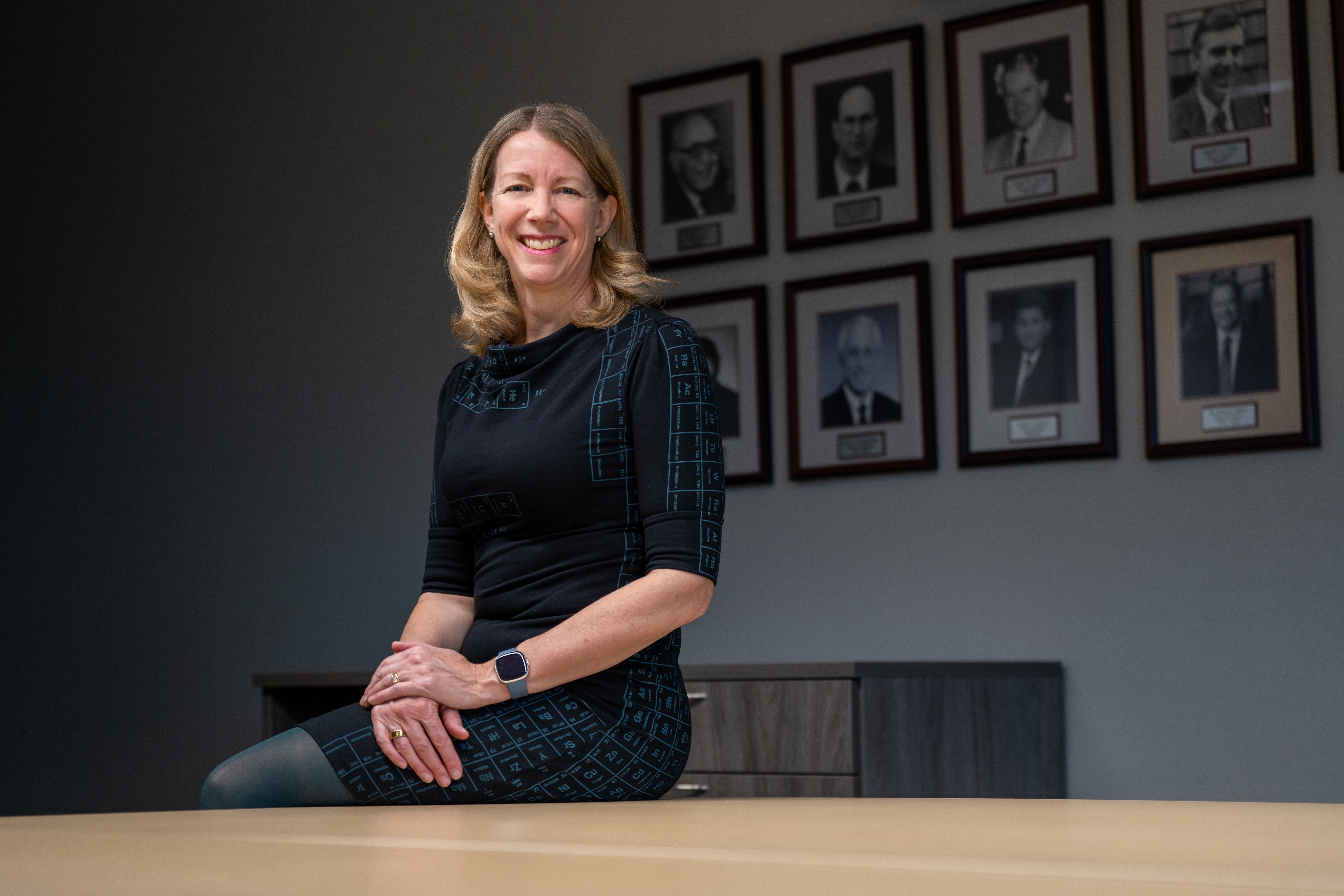 Maureen MacDonald in a dark dress with the periodic table printed on it, sits at the edge of a conference table in a meeting room.